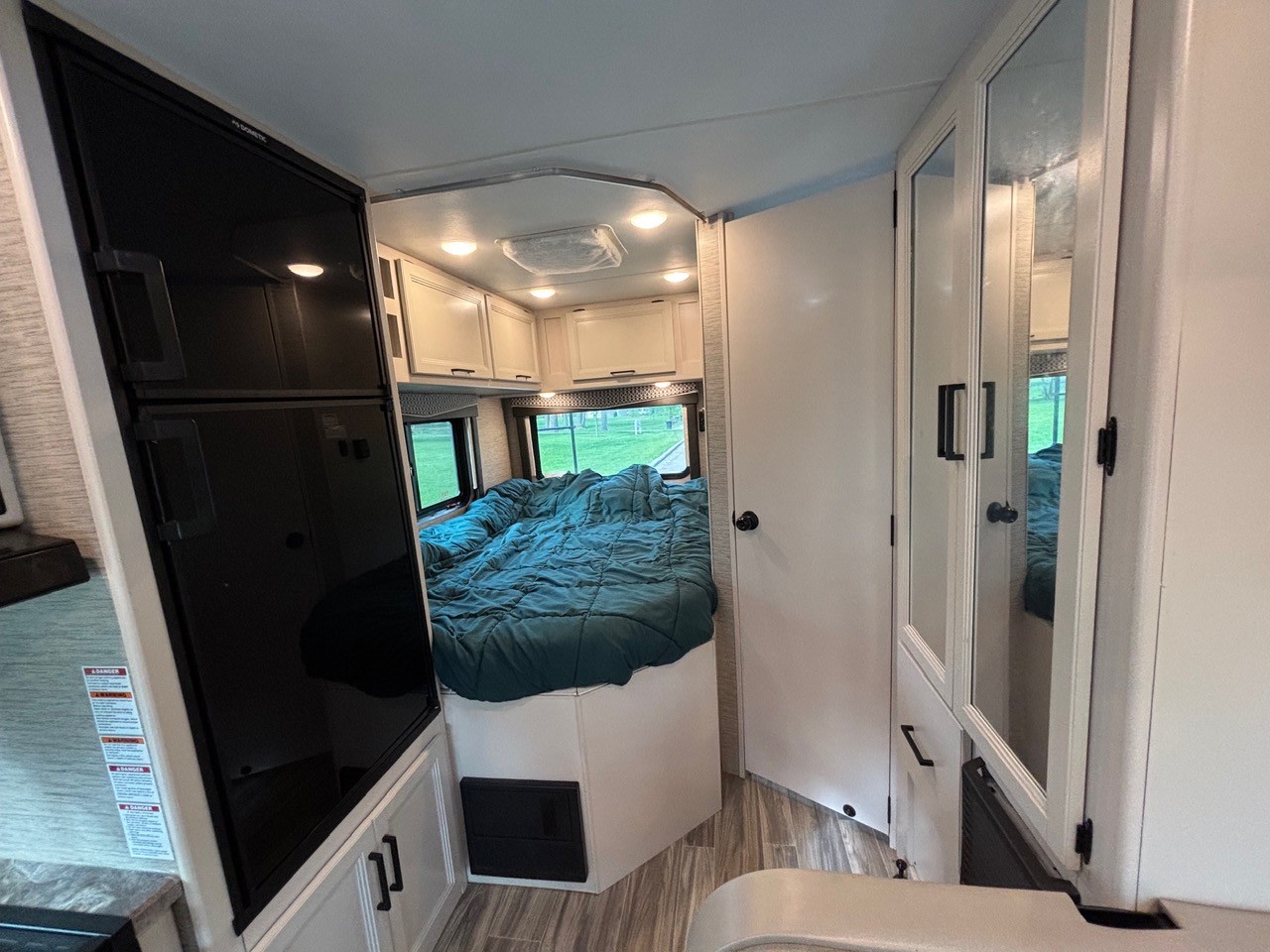 Inside an rv rental: compact bedroom area with black fridge, teal bed by a window, and white mirrored closet.. Thor Four Winds 2023