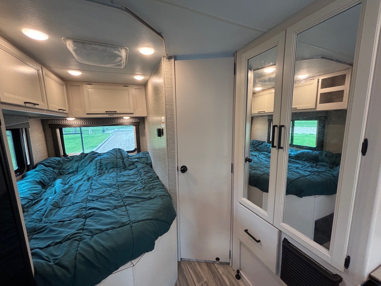 This RV rental bedroom features a comfy teal bed, mirrored wardrobe, overhead cabinets, and a window with outdoor views.. Thor Four Winds 2023