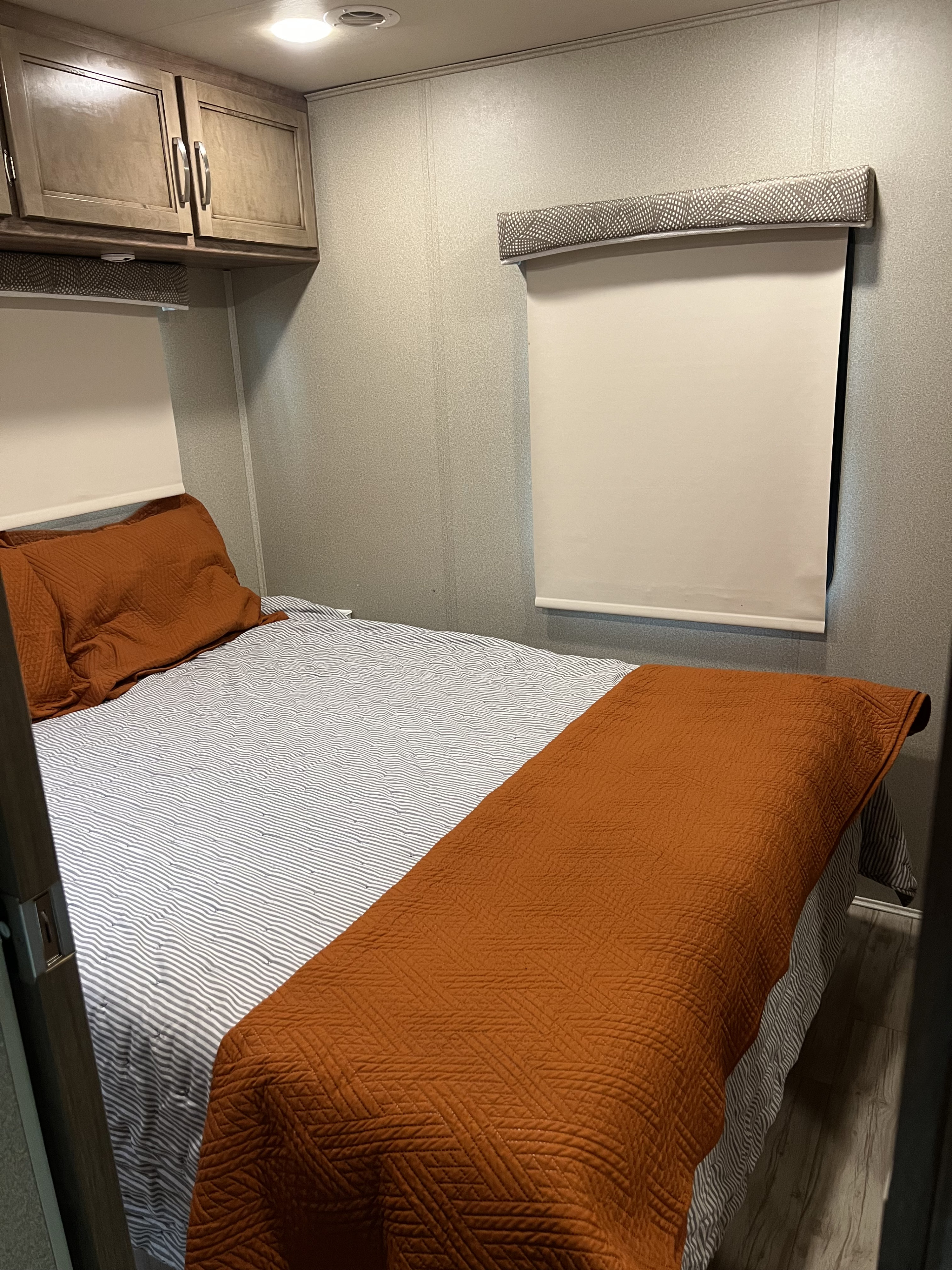Inside an RV rental, this cozy bedroom features a bed with striped sheets, an orange quilt, overhead cabinets, and a roller shade.. Entegra Coach Vision 2019