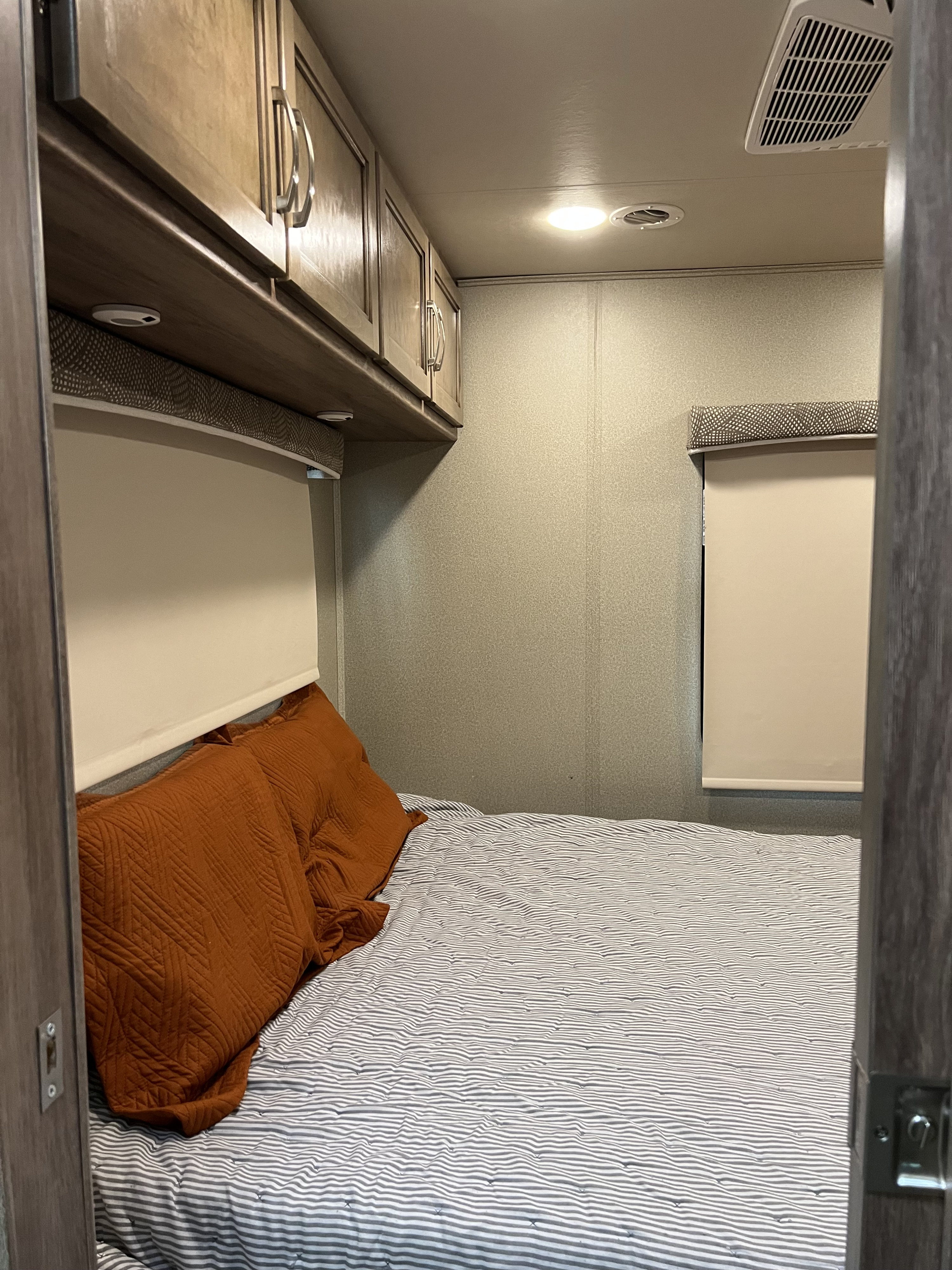 RV rental bedroom view: cozy bed with striped comforter, orange pillows, overhead cabinets, and a window shade for comfortable travel.. Entegra Coach Vision 2019