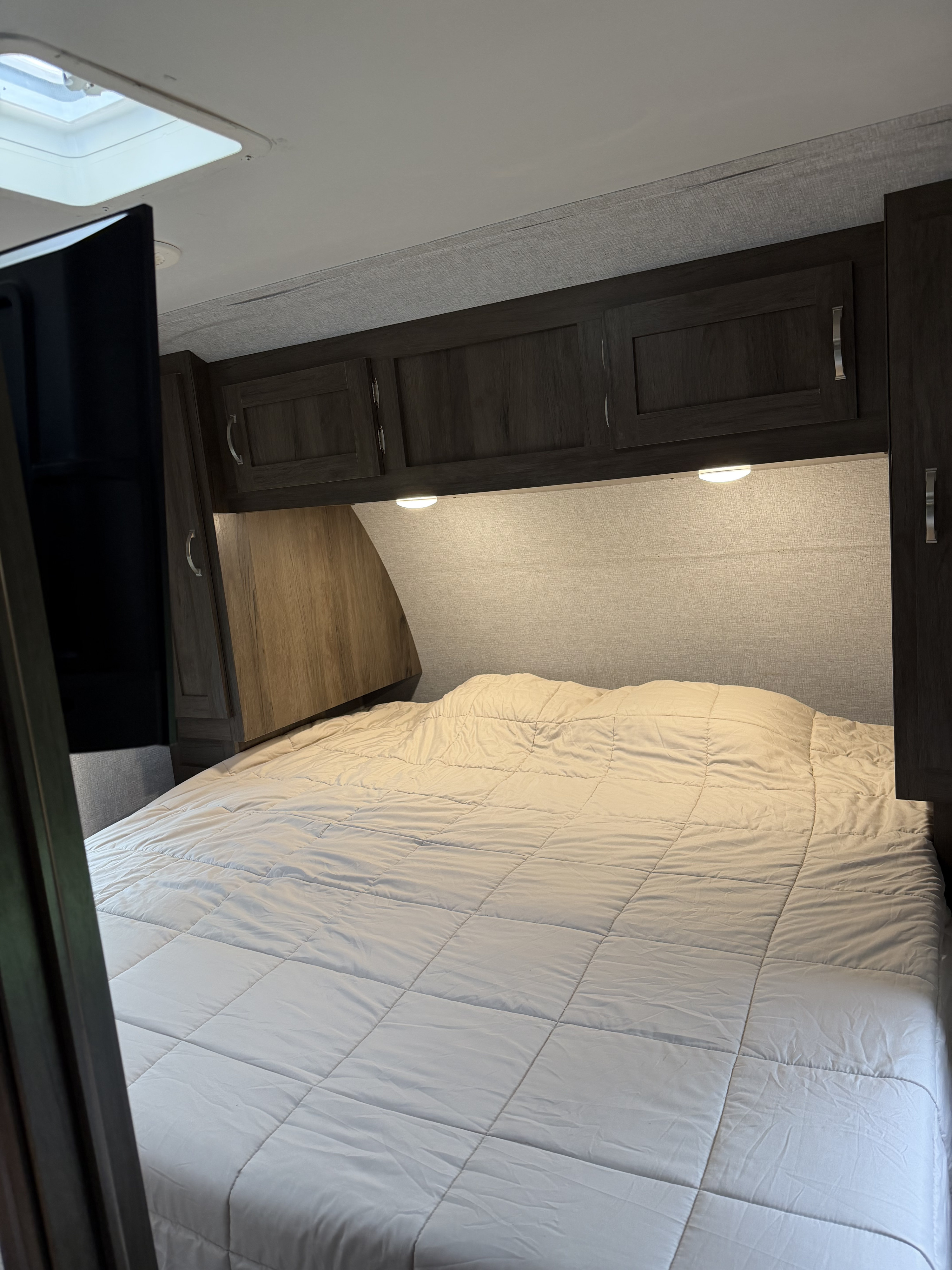 A cozy RV rental bedroom features a neatly made bed, dark overhead cabinets, and a skylight. Perfect for a comfortable stay.. East to West Silver Lake 2021