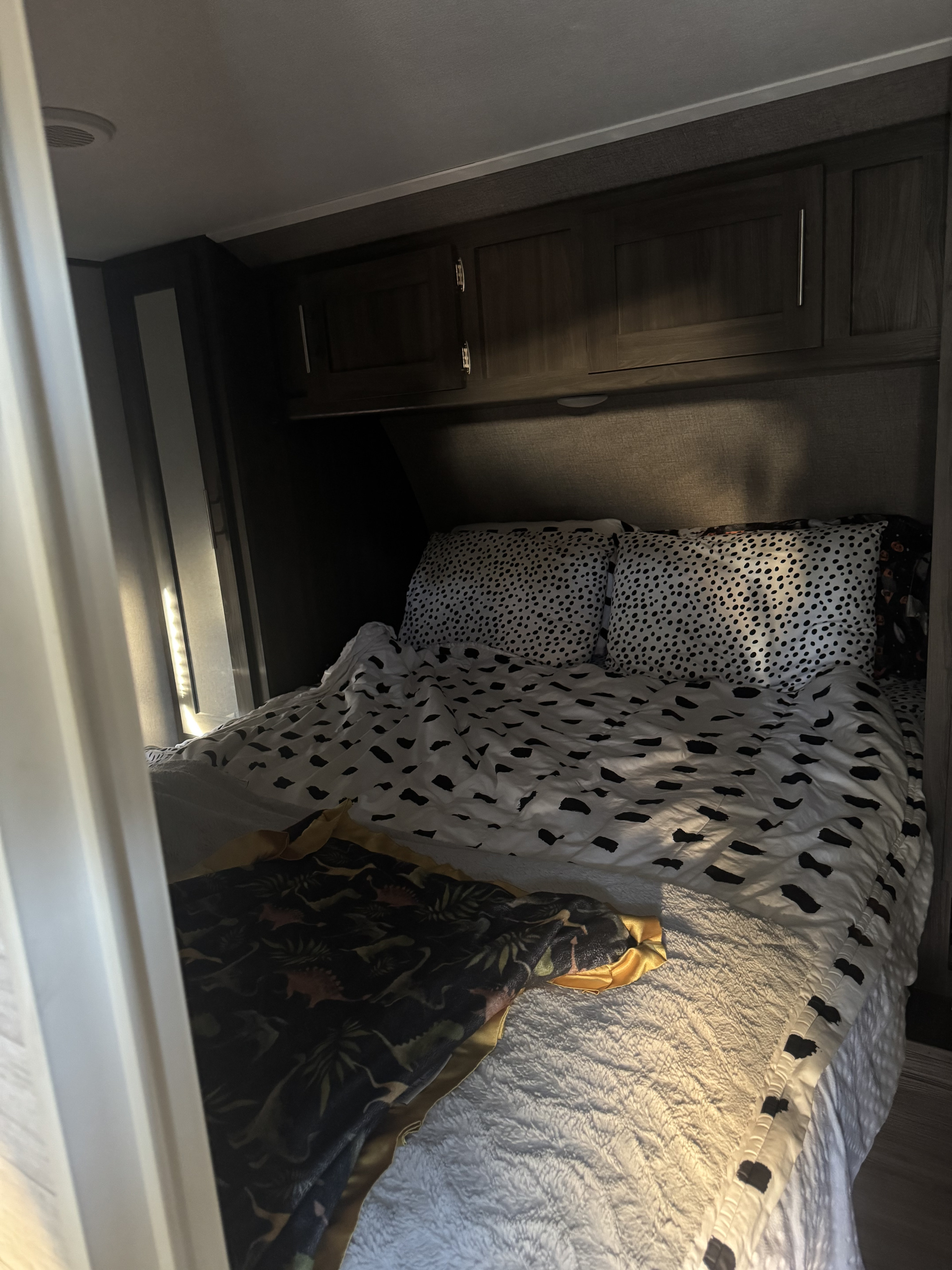 Inside a cozy rv rental: a neatly made bed with spotted pillows, a black-patterned duvet, and a dinosaur blanket under dark wood cabinets.. Forest River Shasta 2021