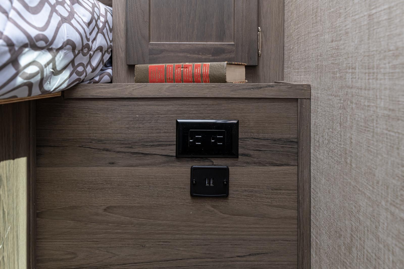 RV rental bedside setup: AC & USB charging outlets on wood paneling, with a book on a shelf, next to a bed.. Gulf Stream Envision 2026