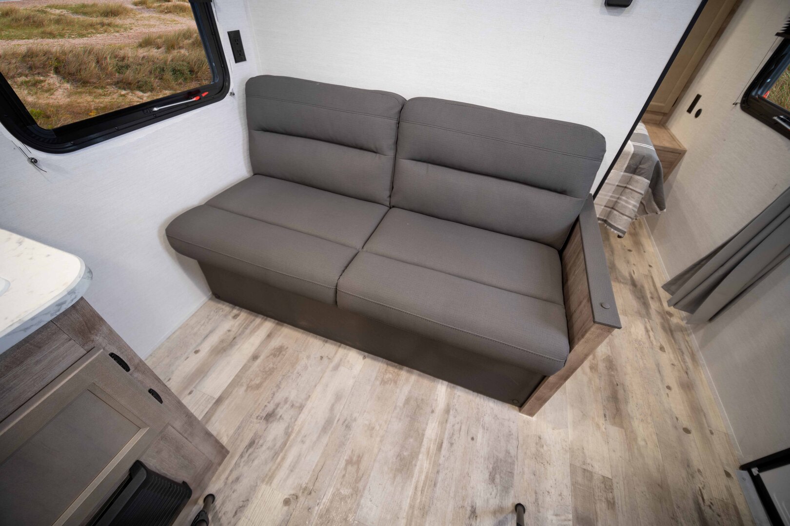 Modern RV rental interior with a compact gray sofa, wood-look floor, and window view of grassy terrain.. Gulf Stream Ameri-Lite 2026