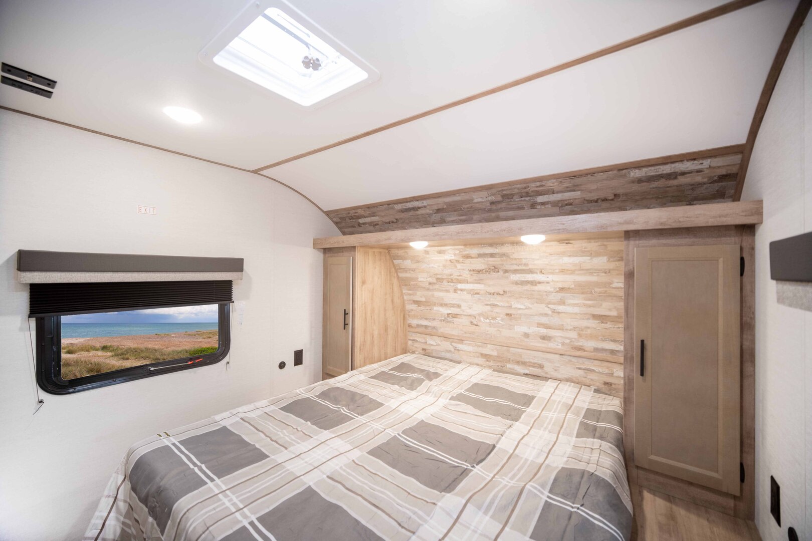 Cozy RV rental bedroom with a plaid bed, ocean view window, skylight, and warm wood-paneled headboard and cabinets.. Gulf Stream Ameri-Lite 2026