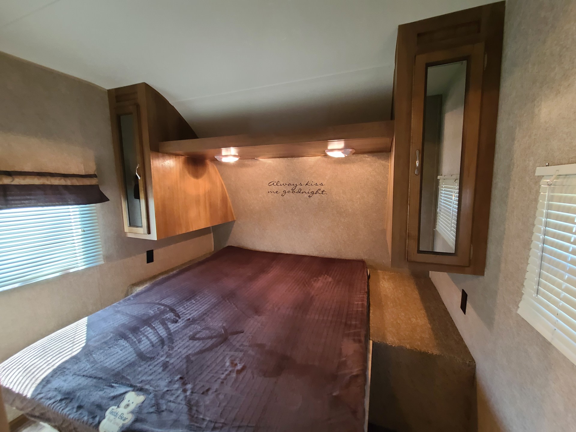 Cozy RV rental bedroom featuring a bed, wooden overhead cabinets, warm lights, and an 'Always kiss me goodnight' wall decal.. Coachmen Catalina 2017