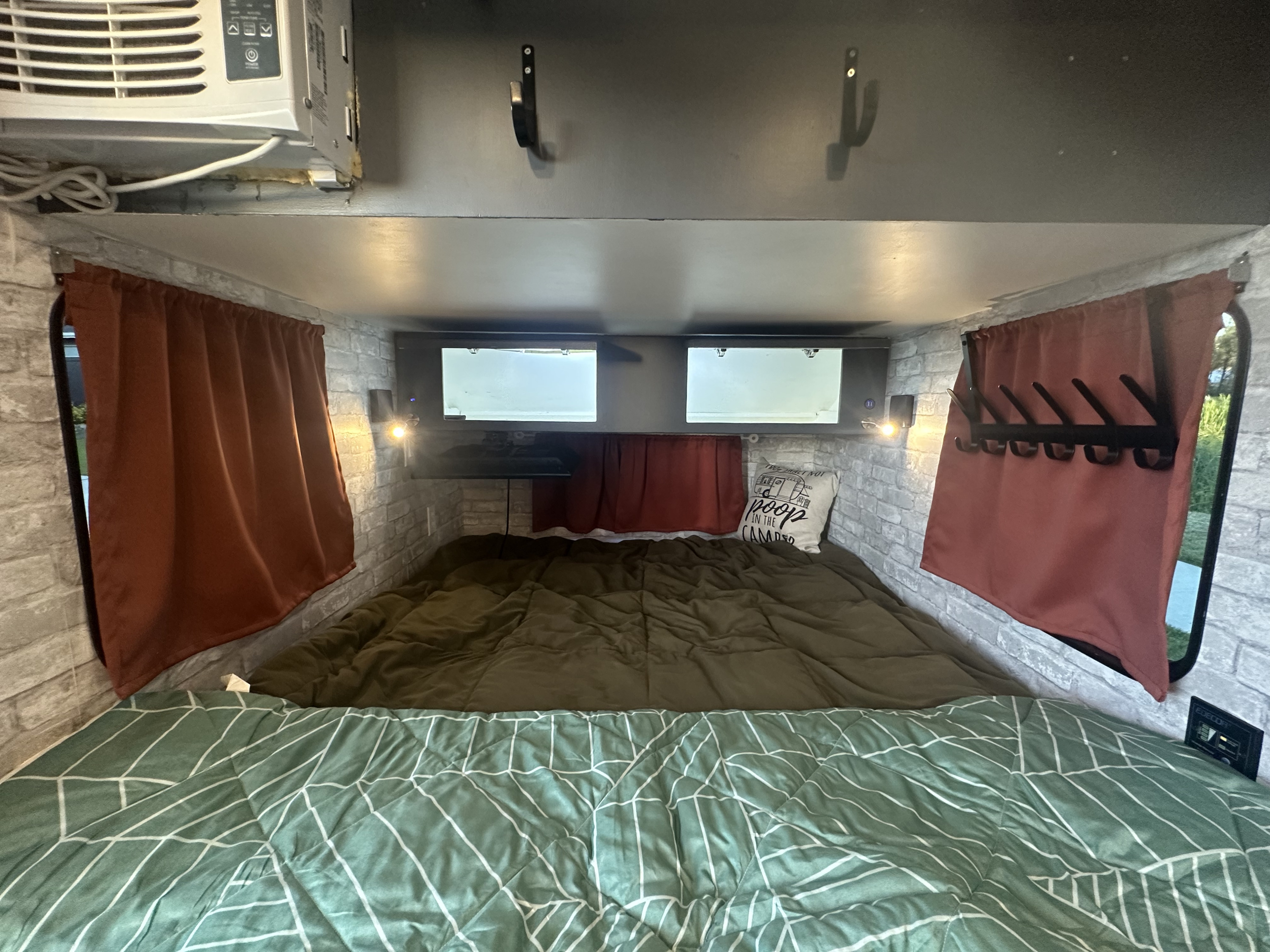 Cozy rv rental sleeping area featuring a bed with patterned bedding, brick-look walls, curtains, and an AC unit for comfort.. Custom Built Standy Karavan 2020