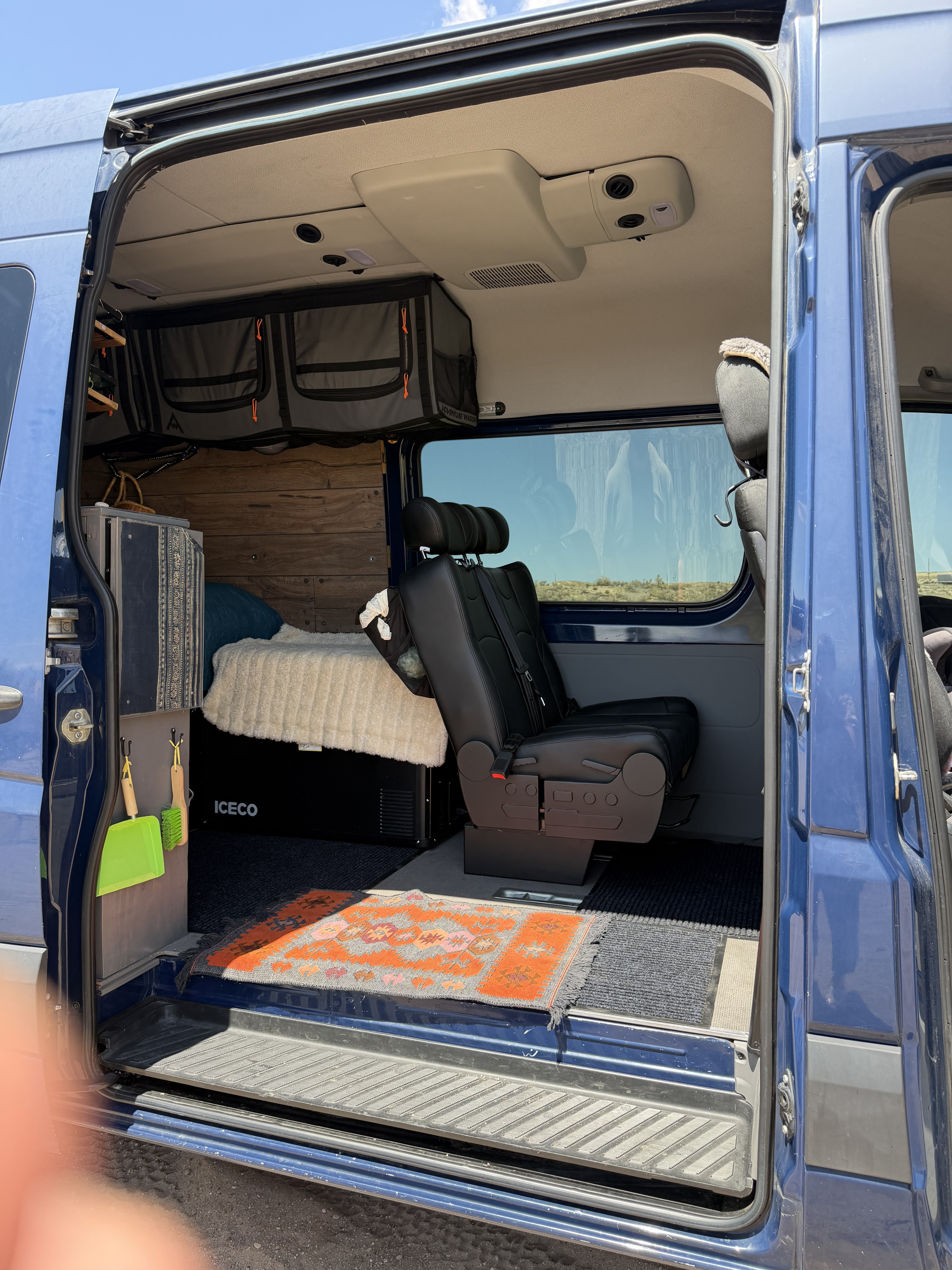 An rv rental camper van interior shows a bed, fridge, passenger seat, custom wood walls, and hanging storage bags.. Mercedes-Benz Sprinter RV Motorhome Campervan 2016