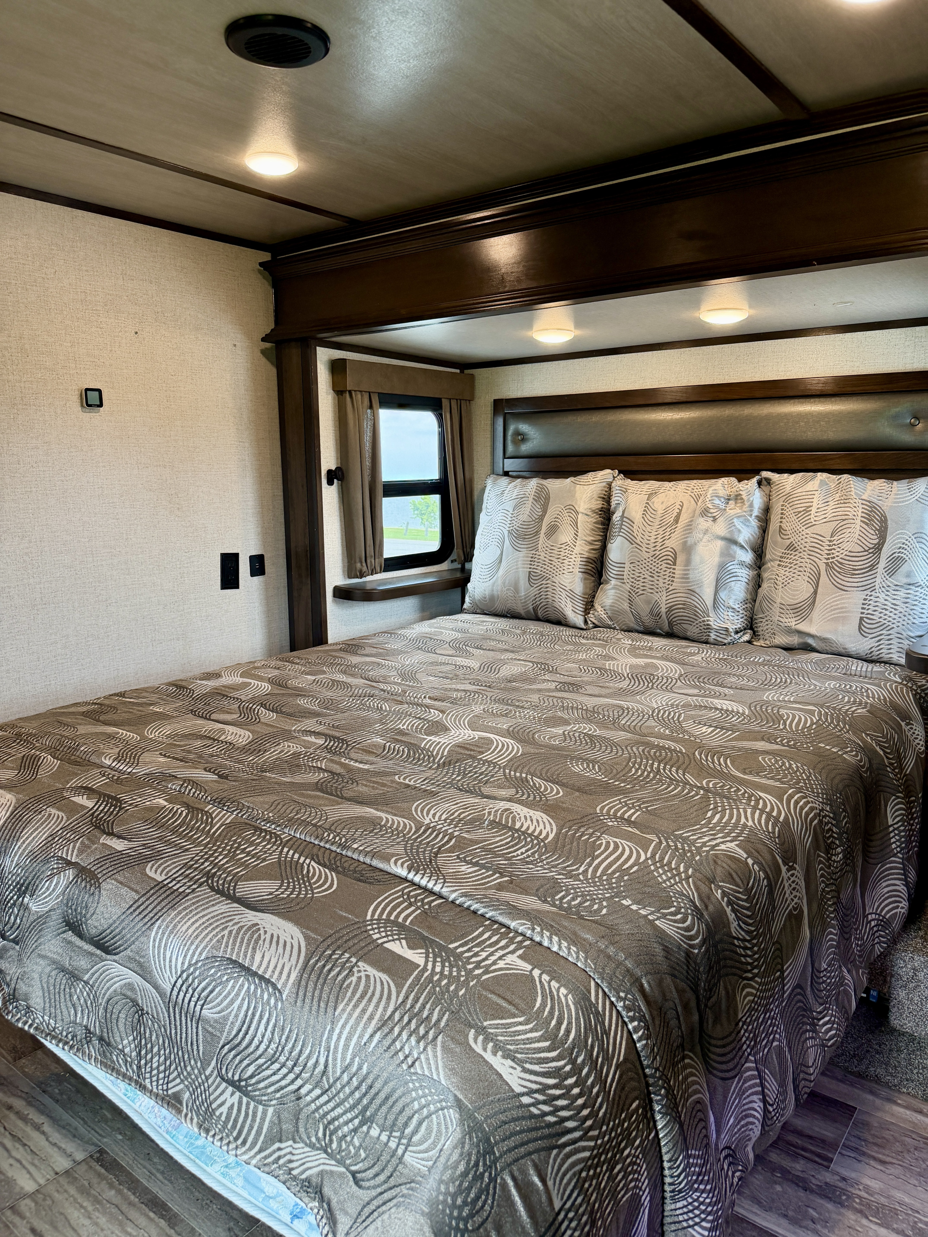 Cozy RV rental bedroom featuring a large bed with patterned bedding, headboard, window, and modern overhead lighting.. Dutchmen Yukon 2021