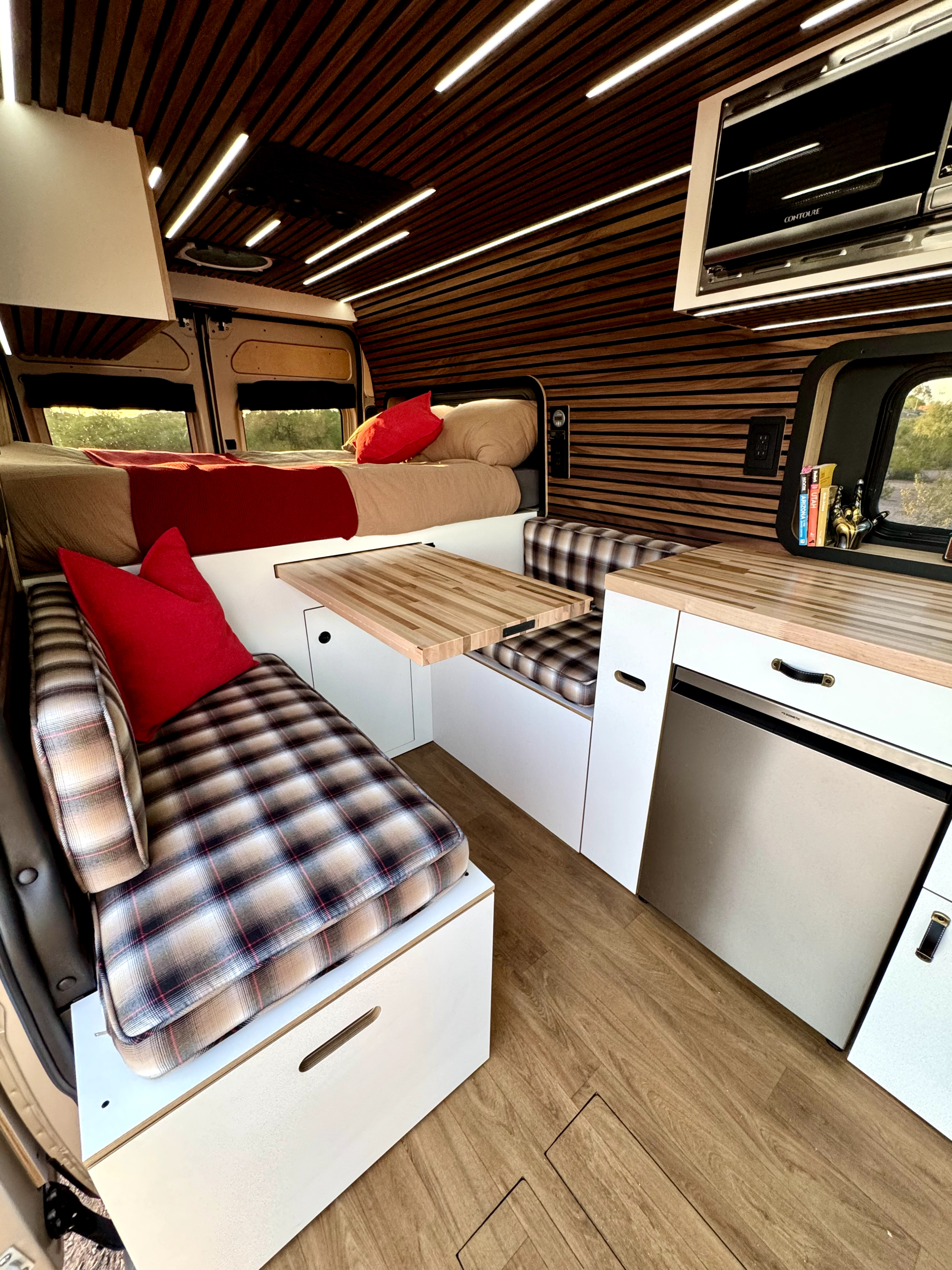 Inviting rv rental interior: compact bed, plaid benches, wooden table, and fridge. Wood-slat ceiling with strip lights adds modern warmth.. Ram Promaster 2500 High Roof 2023