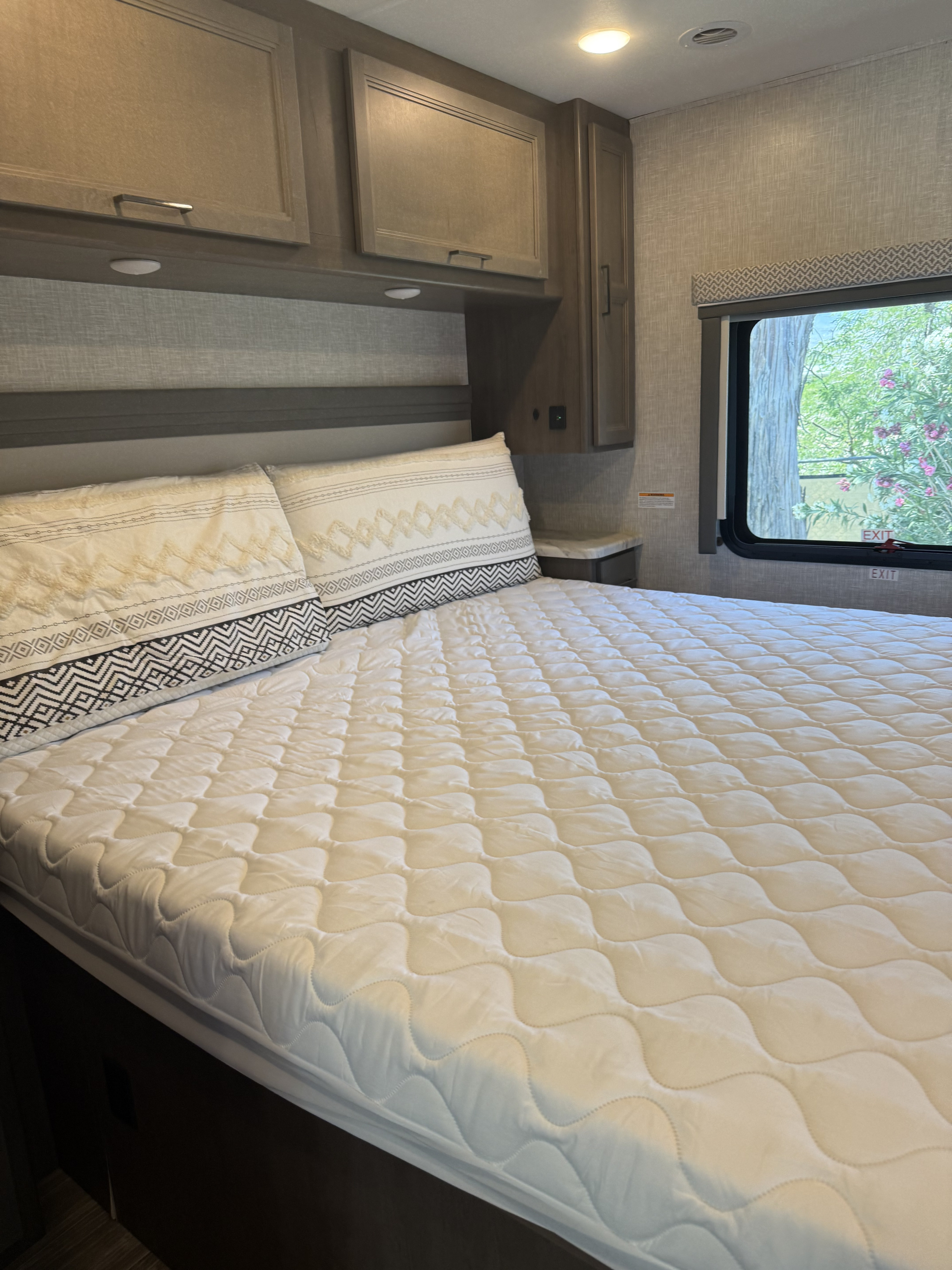 Cozy RV rental bedroom featuring a quilted bed with decorative pillows, overhead cabinets, and a bright window with a nature view.. Thor Hurricane 2024