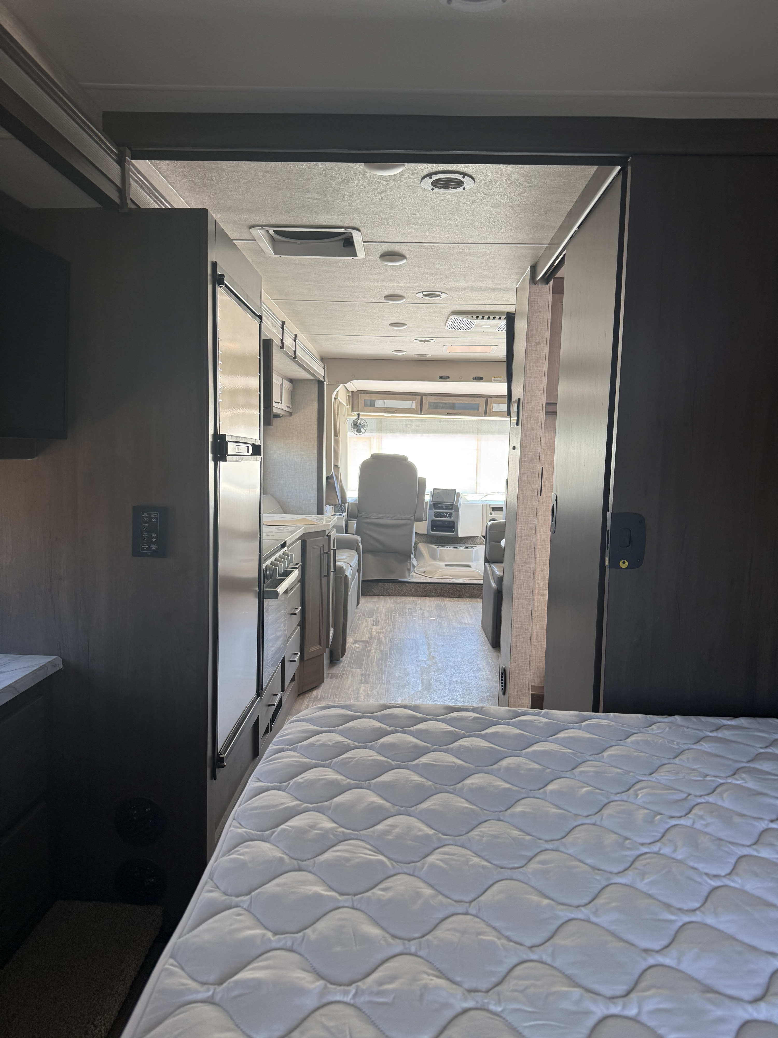View from an RV rental bedroom showing a plush mattress, leading to a kitchen, and the driver's cockpit with seats and dashboard.. Thor Hurricane 2024