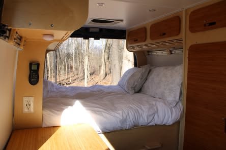 Inside an RV rental: a cozy bed with white linens, wooden cabinets, and a scenic forest view through the large window of this campervan.. Dodge Ram 1500 2022