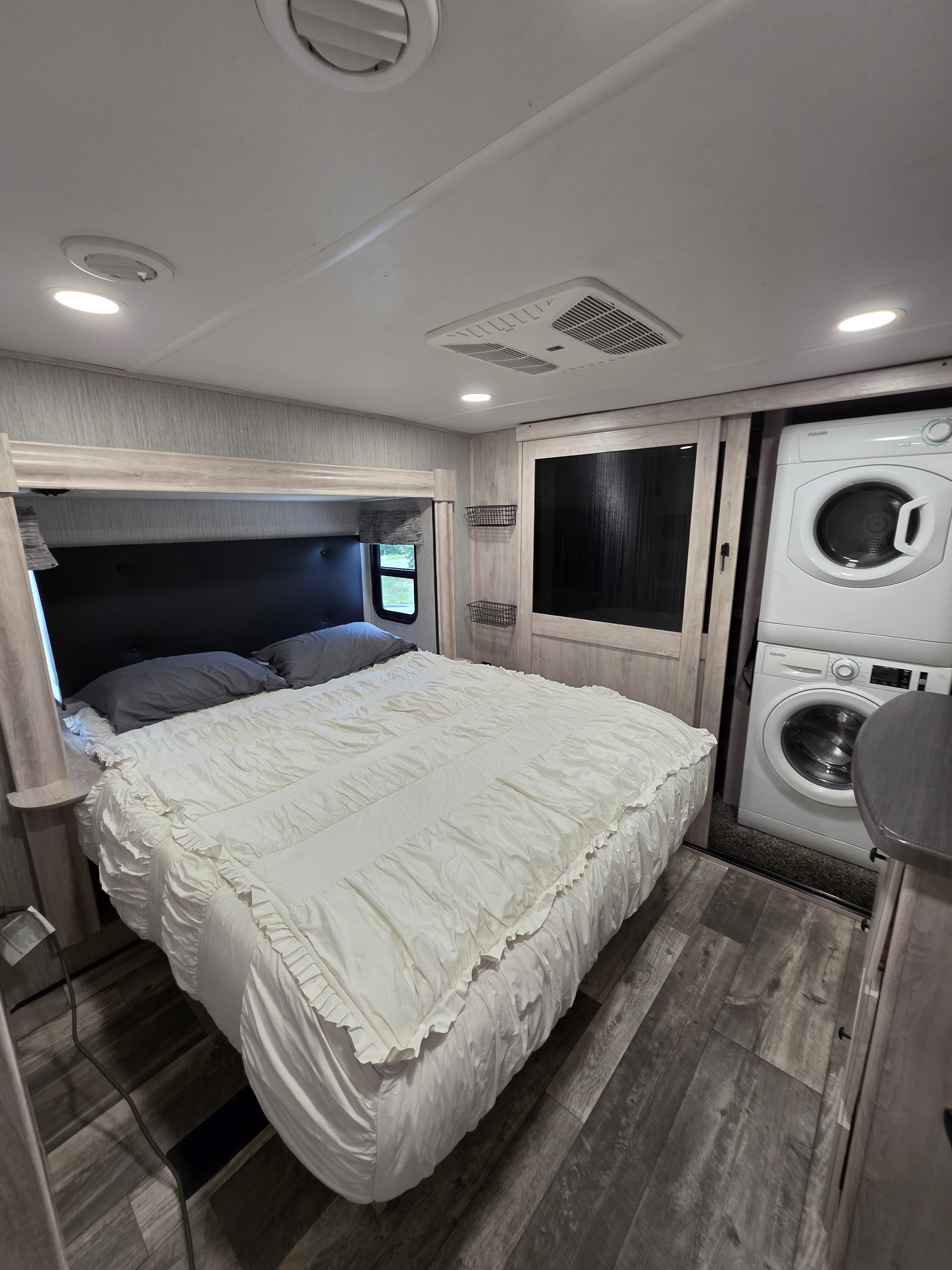 RV rental bedroom with white ruffled bed, gray pillows, stacked washer/dryer, ceiling AC, and recessed lighting.. Forest River Sandpiper 2022