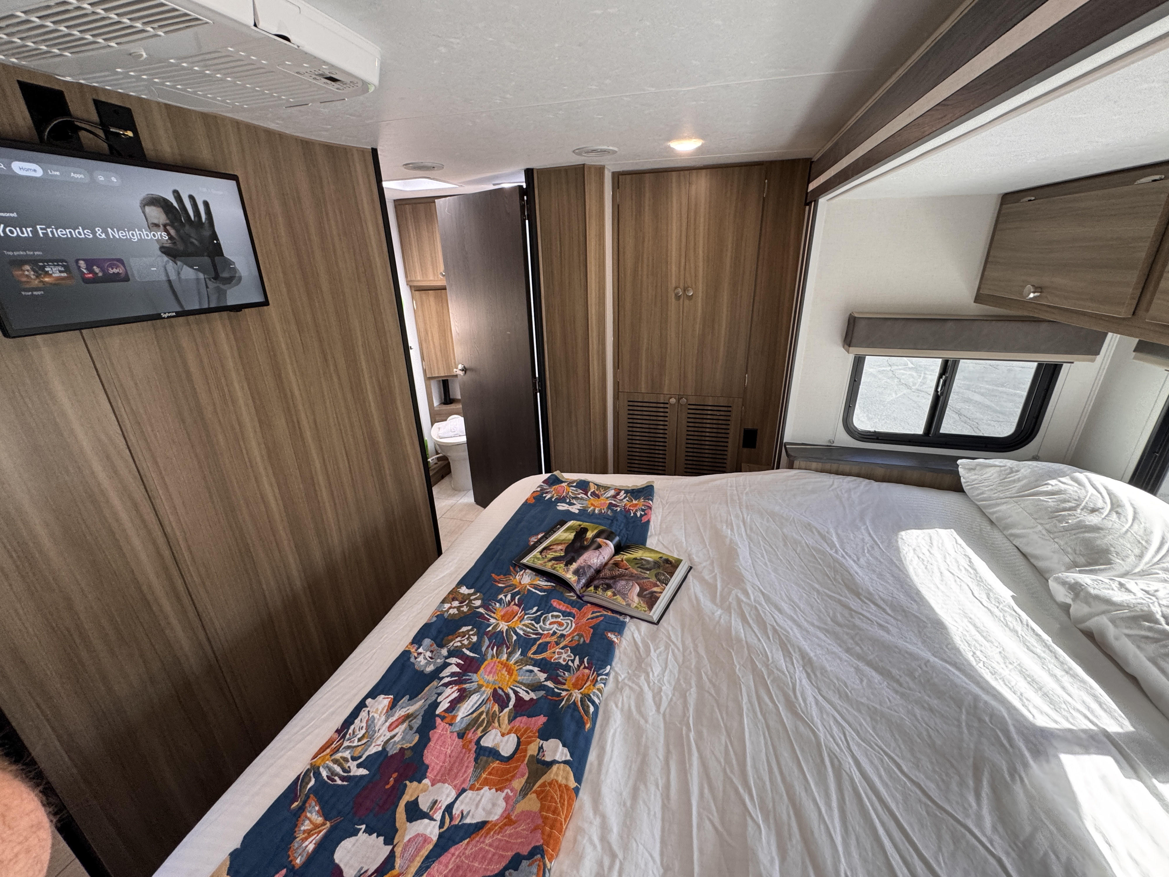 Cozy RV rental bedroom featuring a bed with books, TV, wooden walls, and a partial view of the bathroom.. Forest River Coachmen Freelander 2026