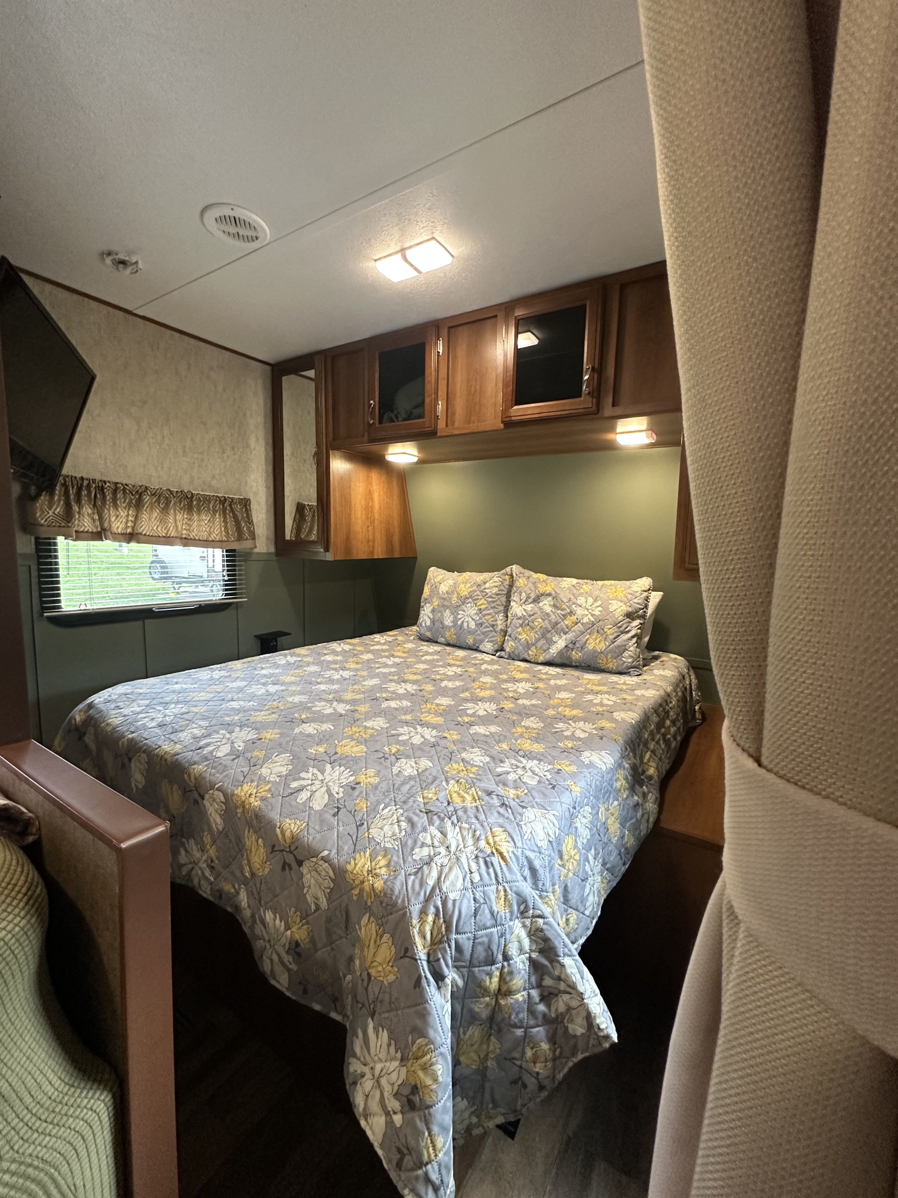 Cozy RV rental bedroom featuring a bed with a grey and yellow floral quilt, overhead cabinets, and a window with blinds.. Avenger Avenger Trailer 2016