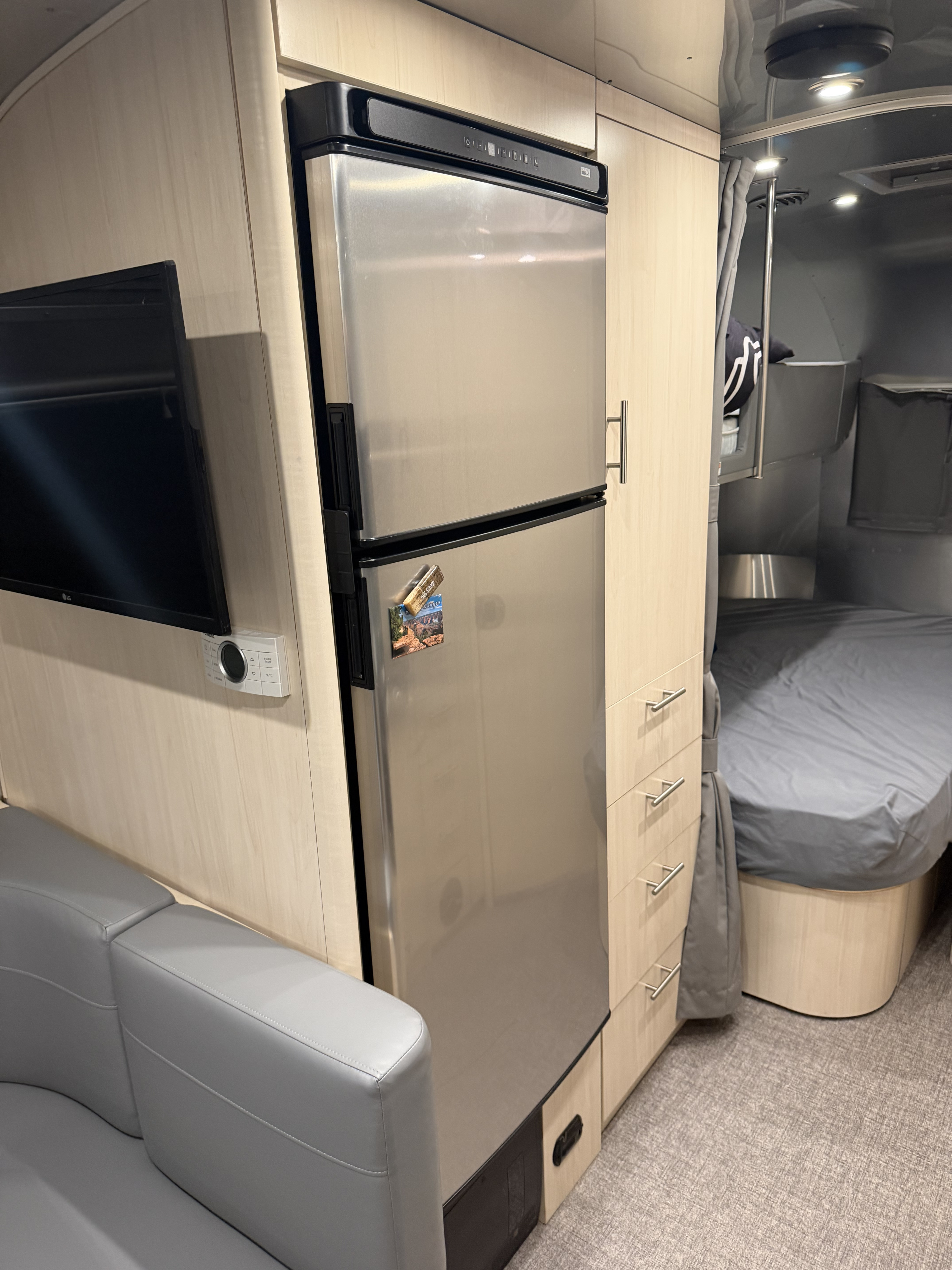 RV rental interior: Stainless steel fridge, TV, controls, light wood cabinetry, gray seating, and a bed area.. Airstream Flying Cloud 2022