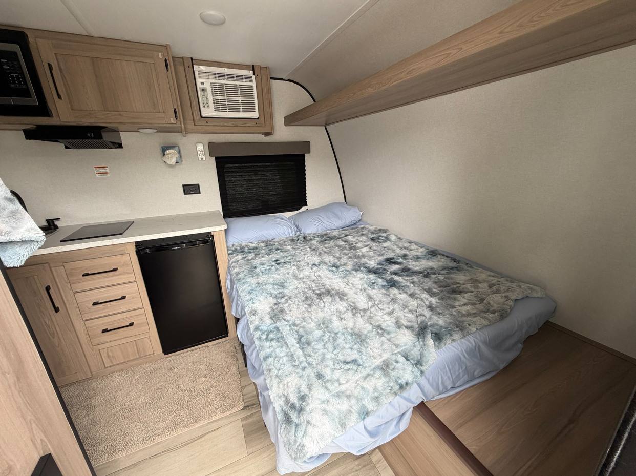Cozy rv rental interior: compact kitchen with microwave, stove, and fridge next to a comfy bed with fluffy blankets.. Keystone RV Coleman Legacy 2026