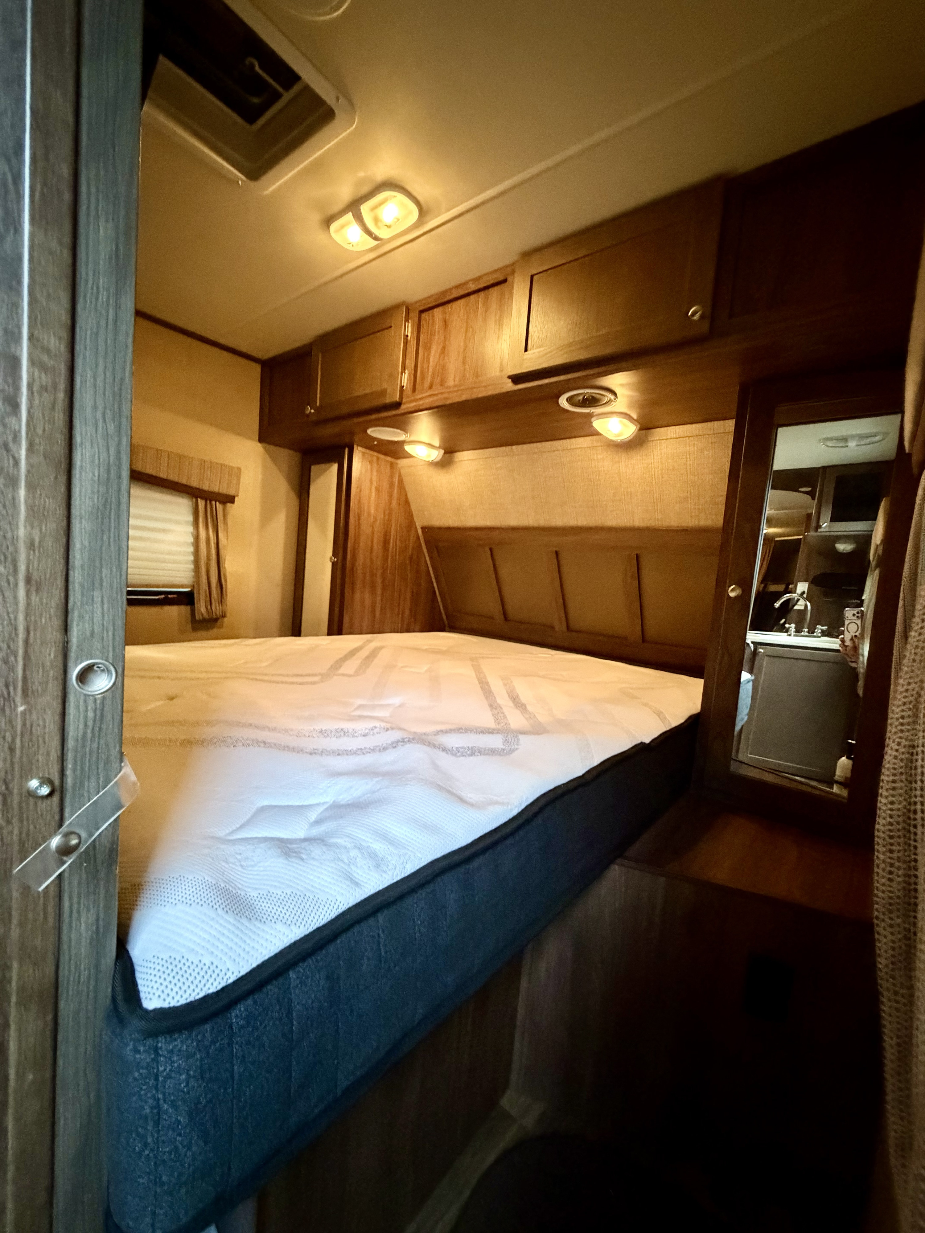 Compact rv rental bedroom featuring a white mattress, wood cabinetry, warm lighting, and a mirror reflecting a kitchenette.. Dutchmen Coleman 2015