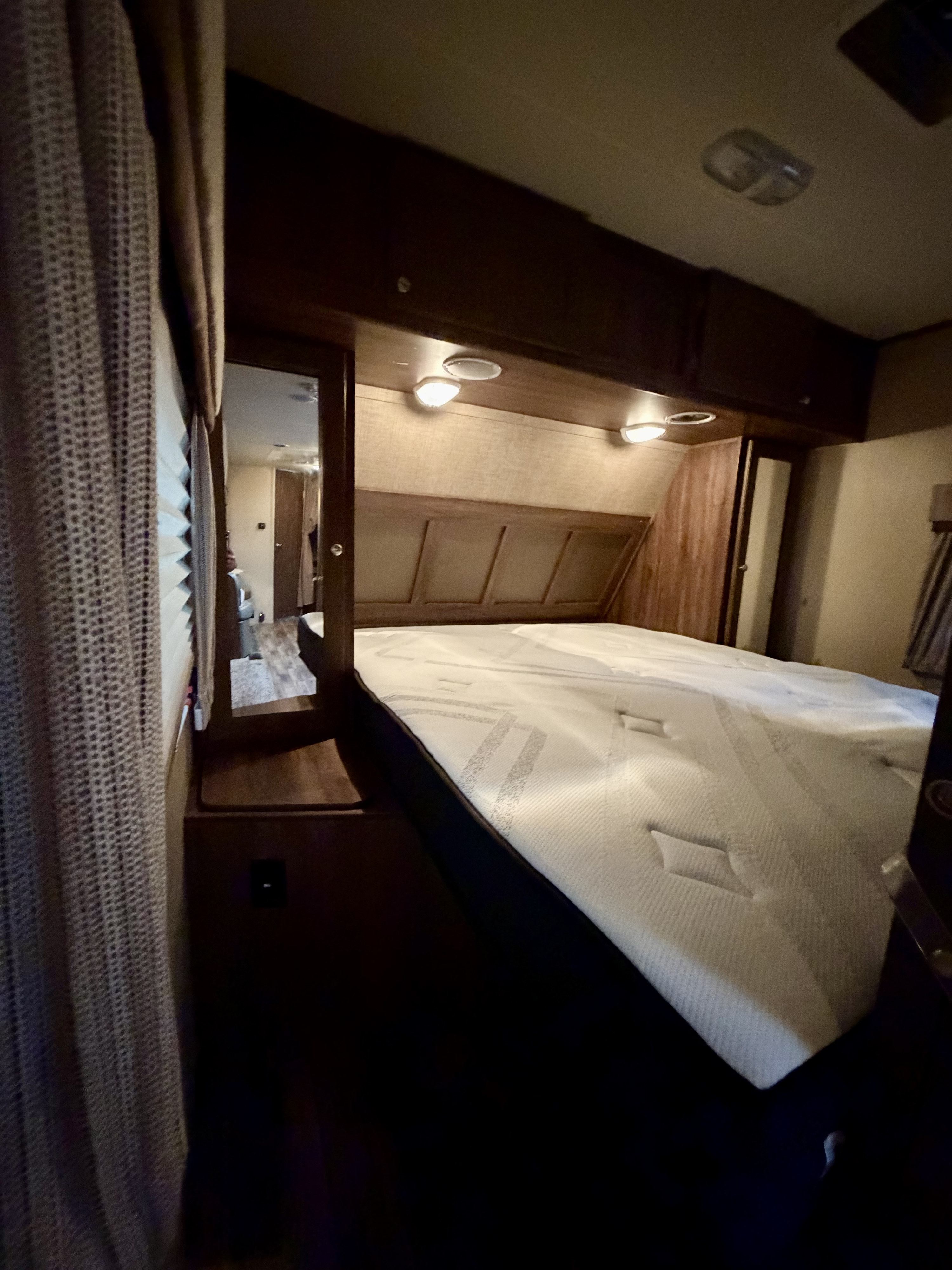 Inside an RV rental, this bedroom features a bed with a white mattress, wooden headboard, overhead cabinets, and mirror.. Dutchmen Coleman 2015