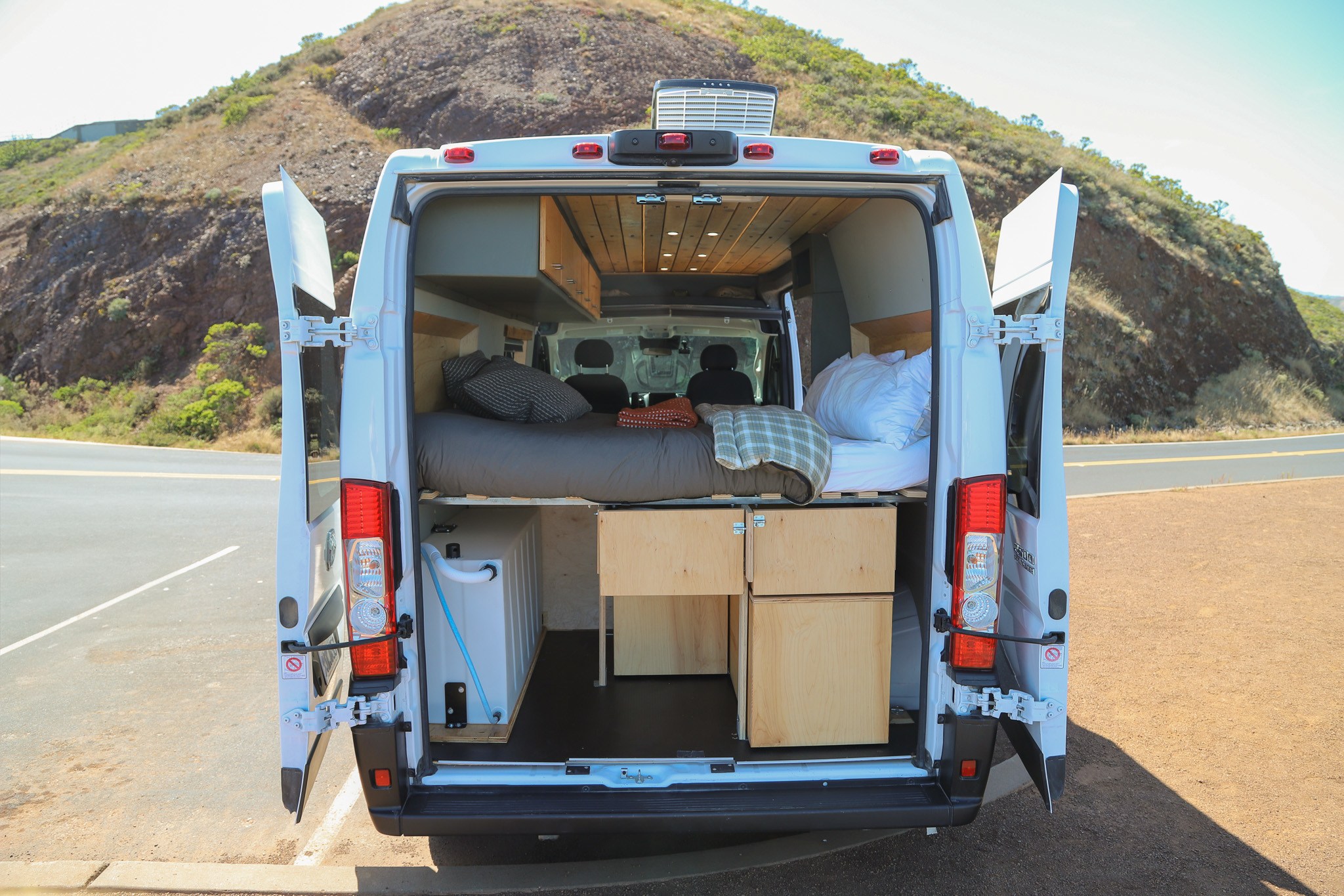 Campervan RV rental: back open, revealing a cozy bed, water tank, and storage. Parked by scenic hills, ready for adventure.. Dodge Ram Promaster 2023