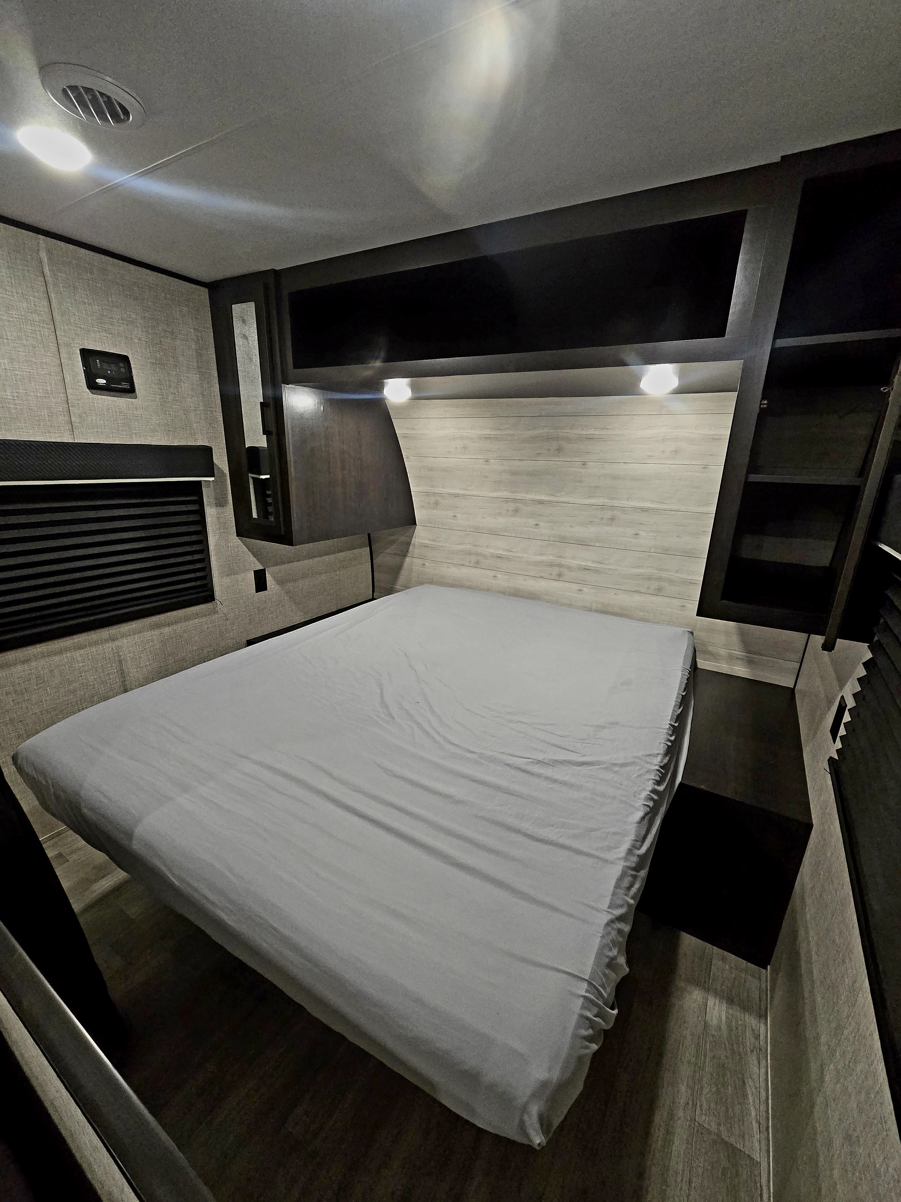 Inside an RV rental: a cozy bedroom featuring a bed with light sheets, wood-look headboard, overhead cabinets, and wall controls.. Jayco Jay Flight 2023
