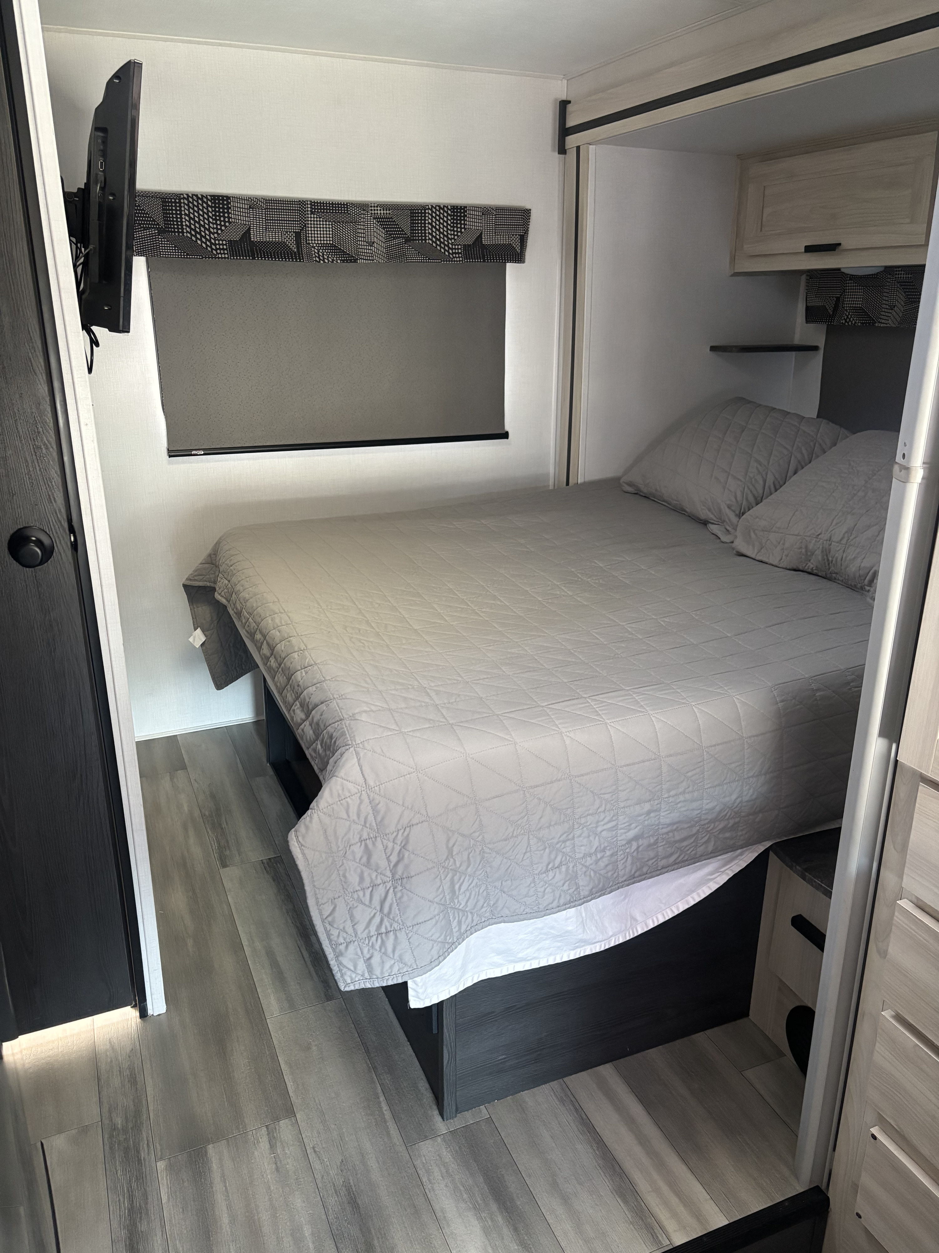 Comfortable RV rental bedroom featuring a made bed with gray quilted bedding, wall-mounted TV, and light wood cabinetry.. Forest River Sunseeker LE 2023