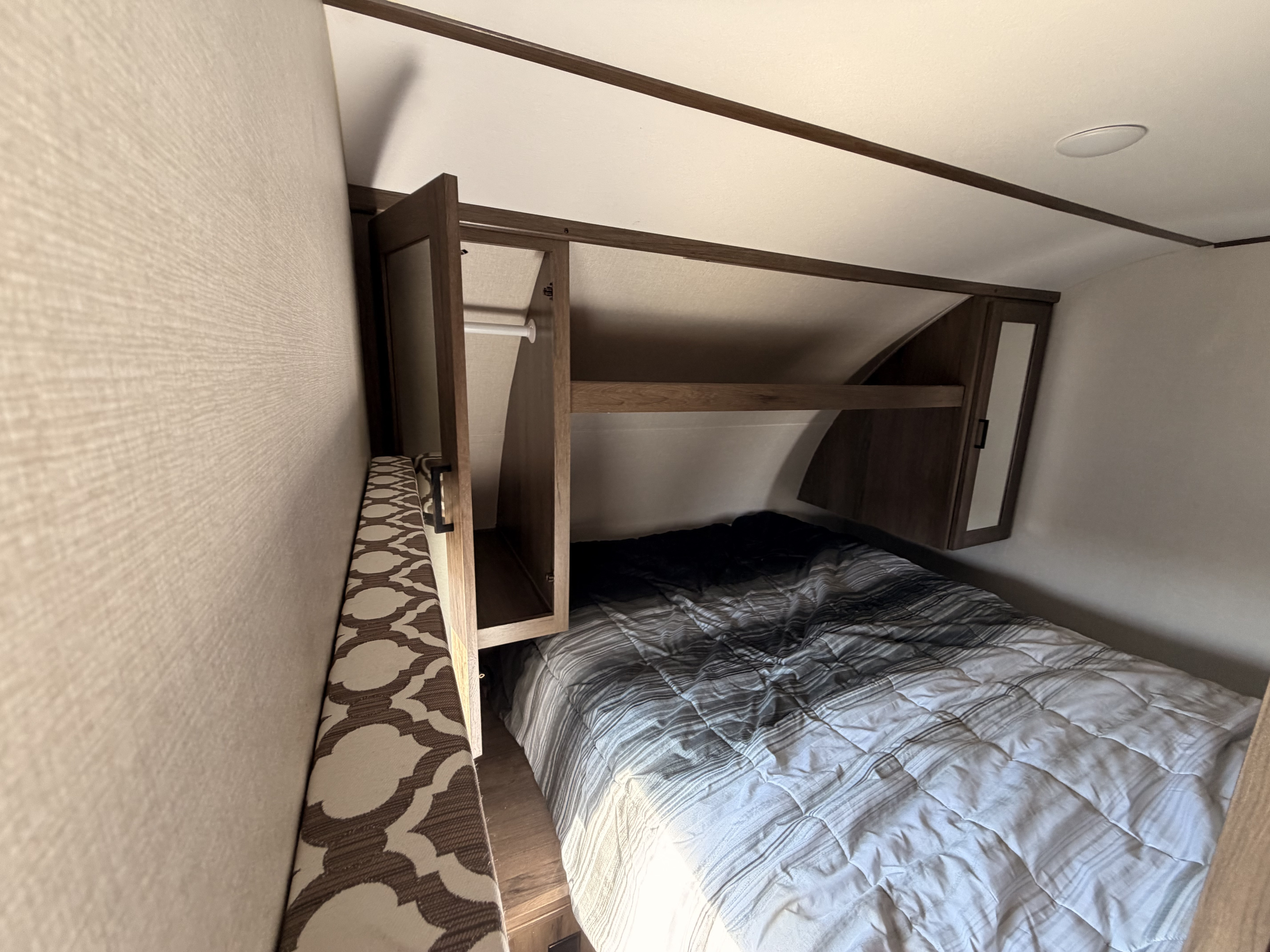 Cozy RV rental bedroom with a striped bed, overhead storage (one open with a hanging rod), and a patterned bench.. Prime Time Tracer 2020