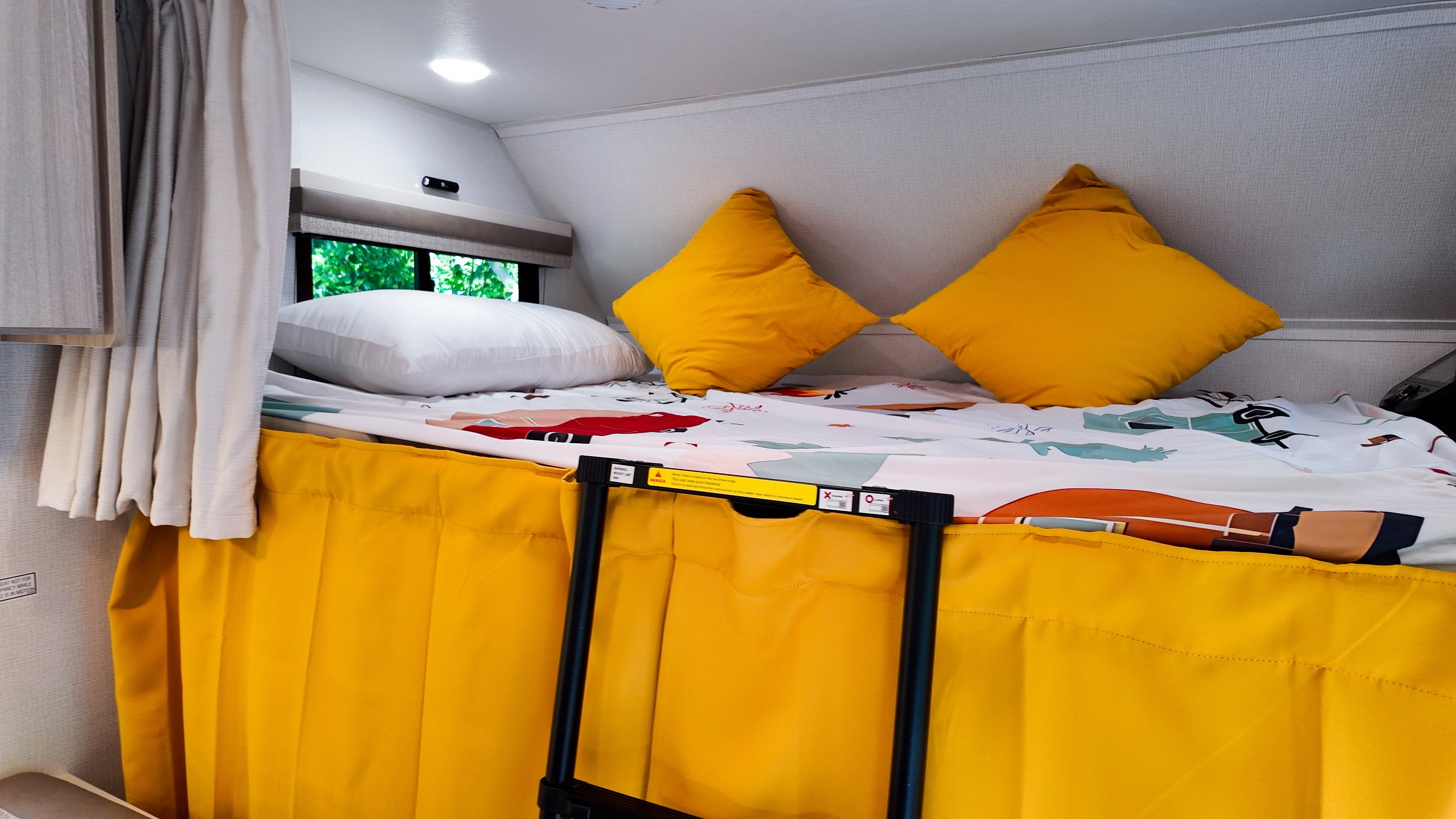 Bright RV rental sleeping area: an elevated bed with yellow pillows, colorful duvet, and a ladder. Green trees outside window.. Forest River Entrada 2026