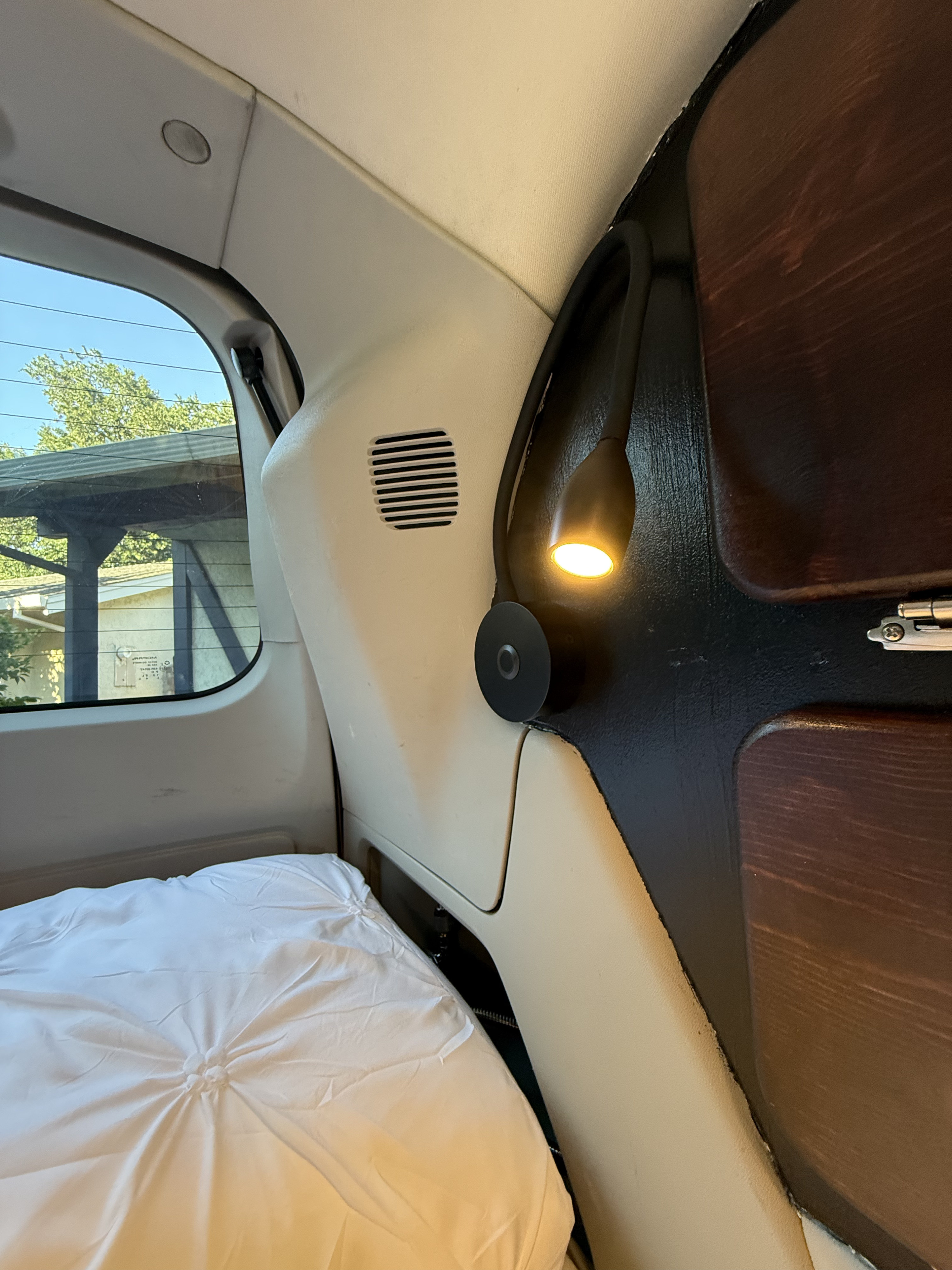 Cozy rv rental interior with a white bed, window view, and a warm reading lamp attached to a dark wood panel.. Chrysler Town & County 2010