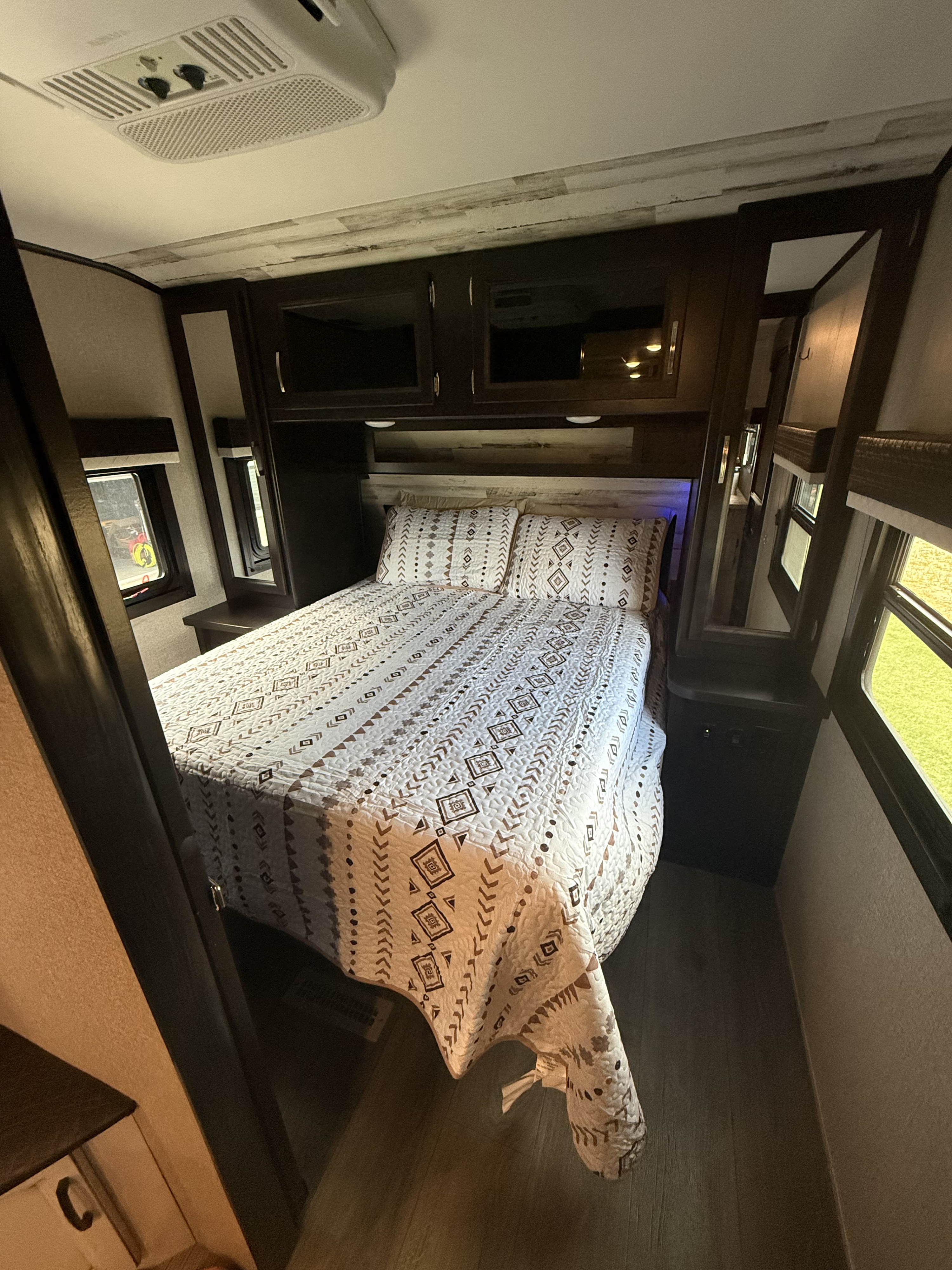 Cozy RV rental bedroom featuring a patterned bed, overhead cabinets, windows, and an AC unit.. Jayco Jay Feather 2021