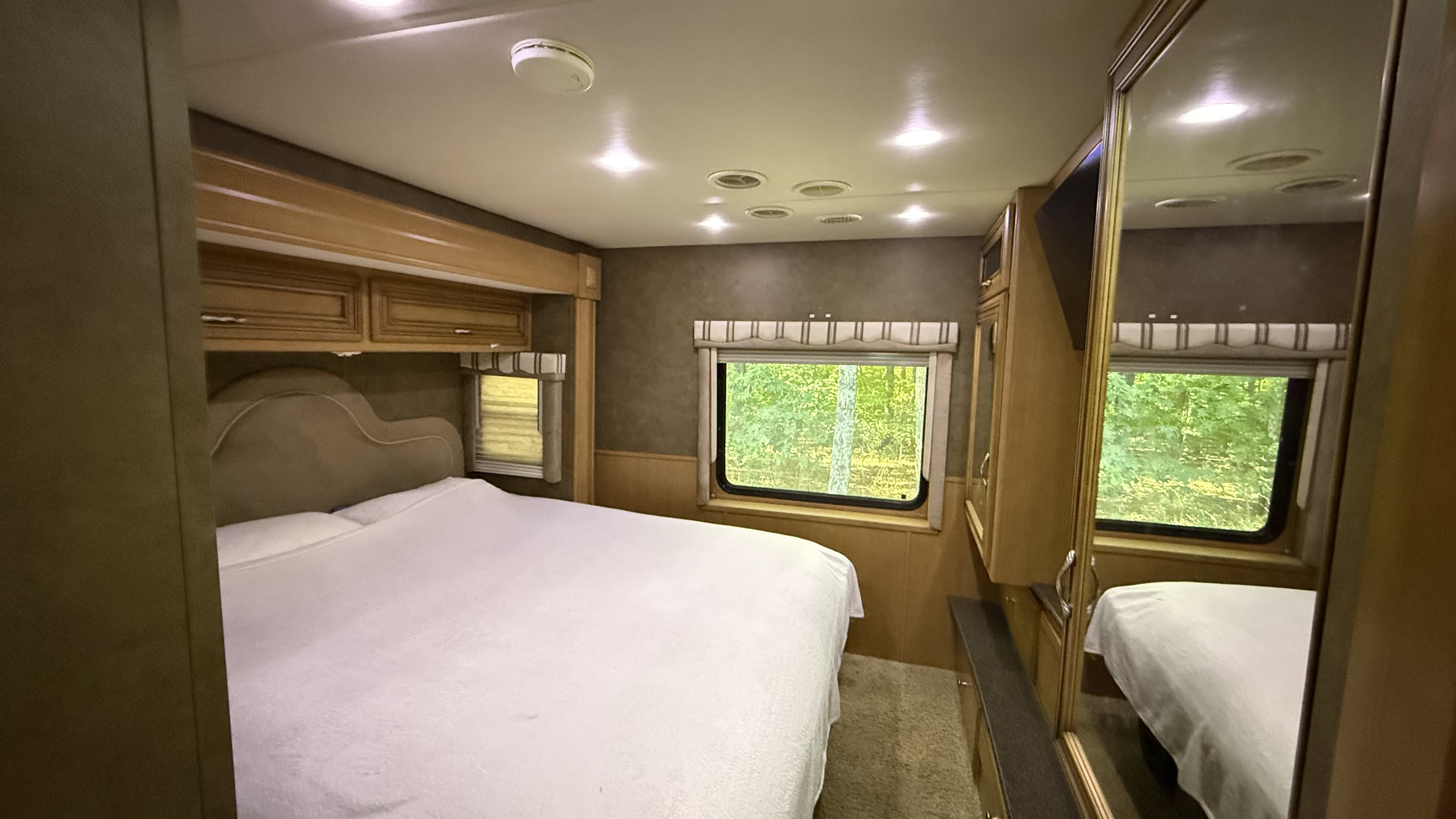 Spacious RV rental bedroom with a large bed, padded headboard, wood cabinets, and windows framing green trees. Bright & clean.. Newmar Baystar 3012 2013