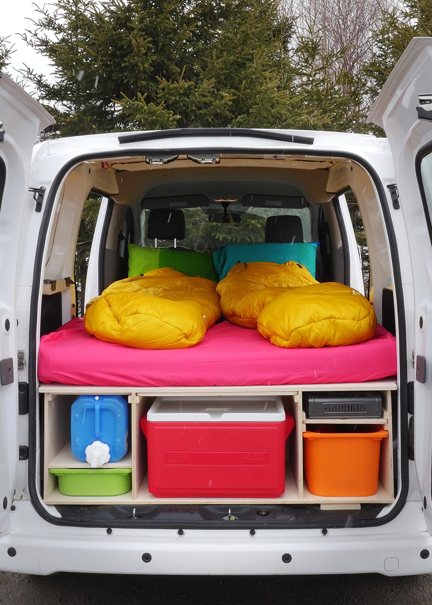 Cozy RV rental setup: van's rear reveals a pink bed, yellow sleeping bags, and organized storage with a cooler, water jug, and stove.. Nissan NV200 2020