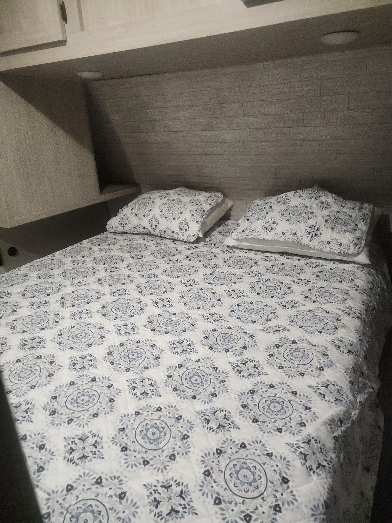 Cozy RV rental bedroom with a white and blue patterned comforter and pillows, framed by light wood cabinets above.. Coachmen 29THS 2025