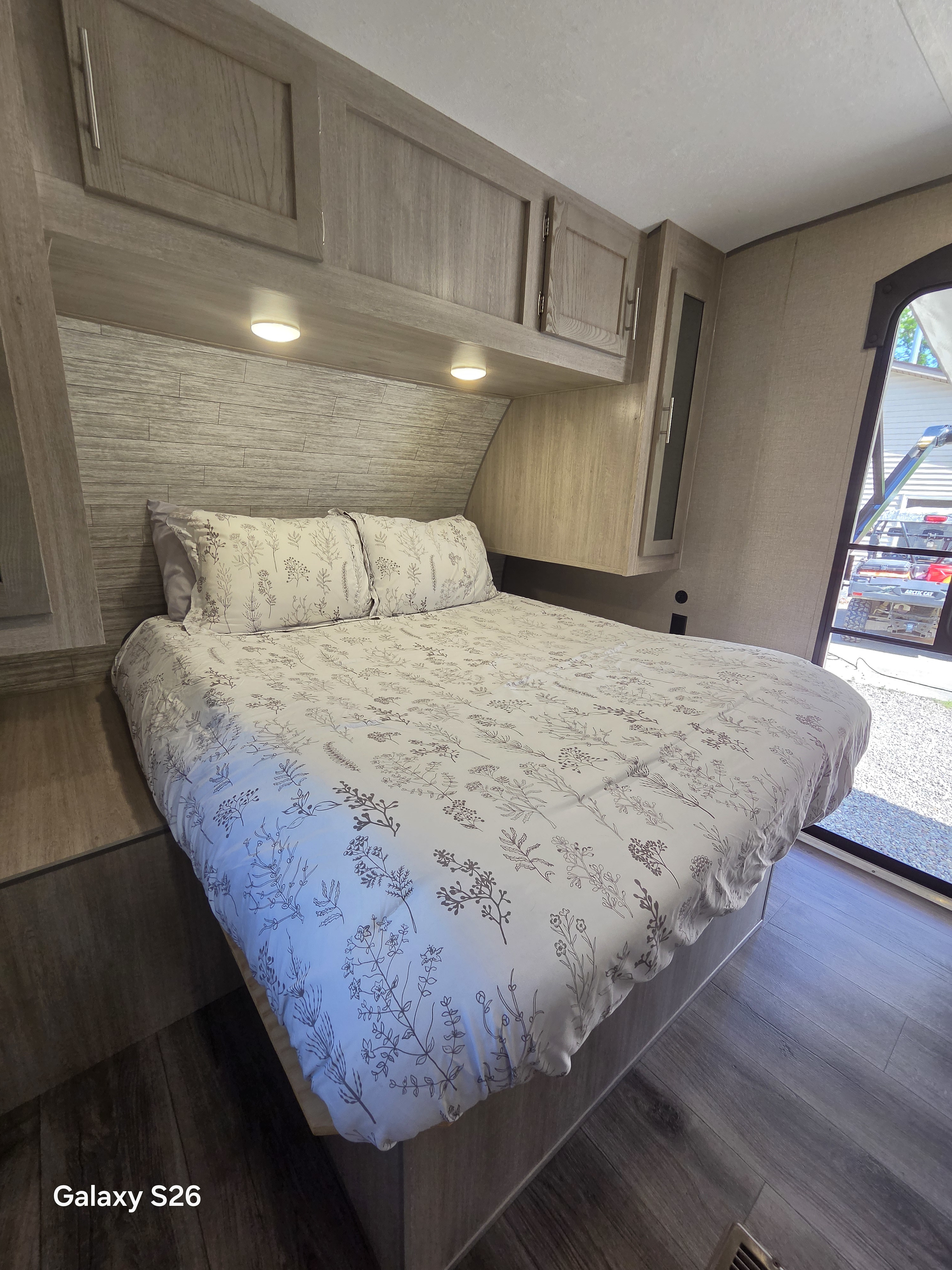 Cozy RV rental bedroom featuring floral bedding, overhead storage, wood paneling, and a view of the outdoors.. Forest River Coachmen Catalina Legacy 2023