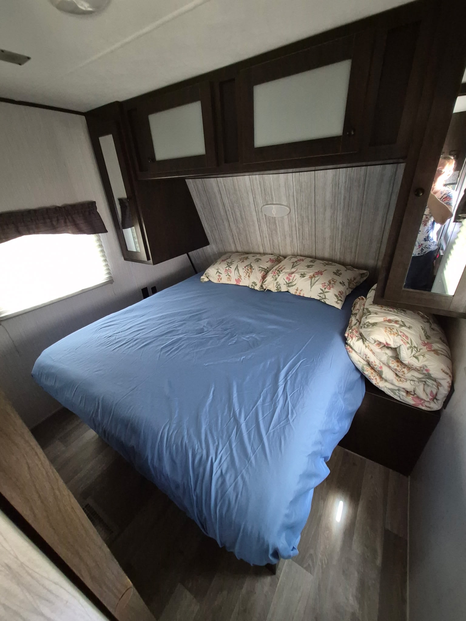 Compact RV rental bedroom: bed with blue sheets, floral pillows/comforter, overhead cabinets, and a window.. Heartland Pioneer 2021