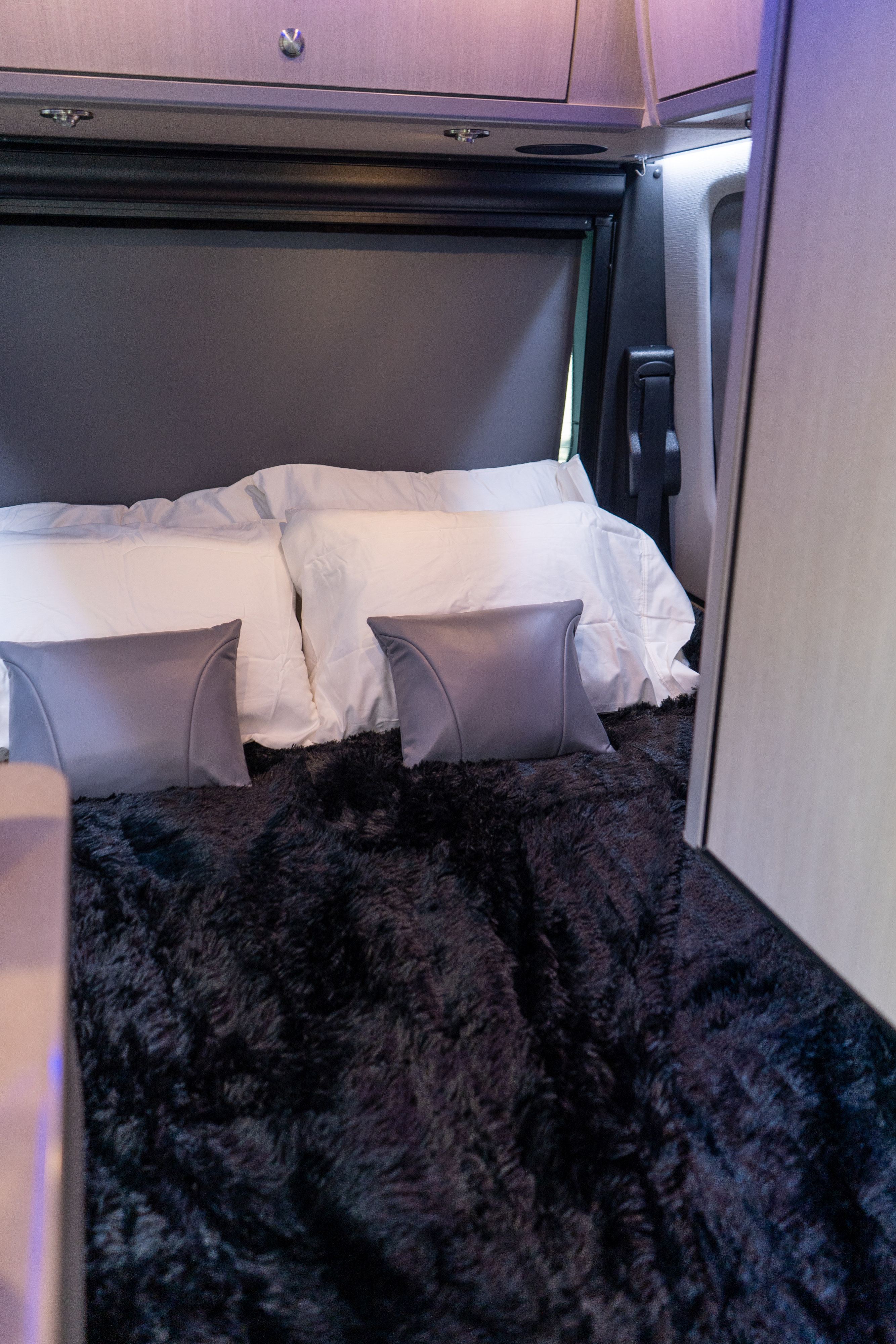 This RV rental's cozy bed features white bedding, grey pillows, and a fluffy black blanket, with overhead storage and lighting.. Grech RV Turismo Ion 2026