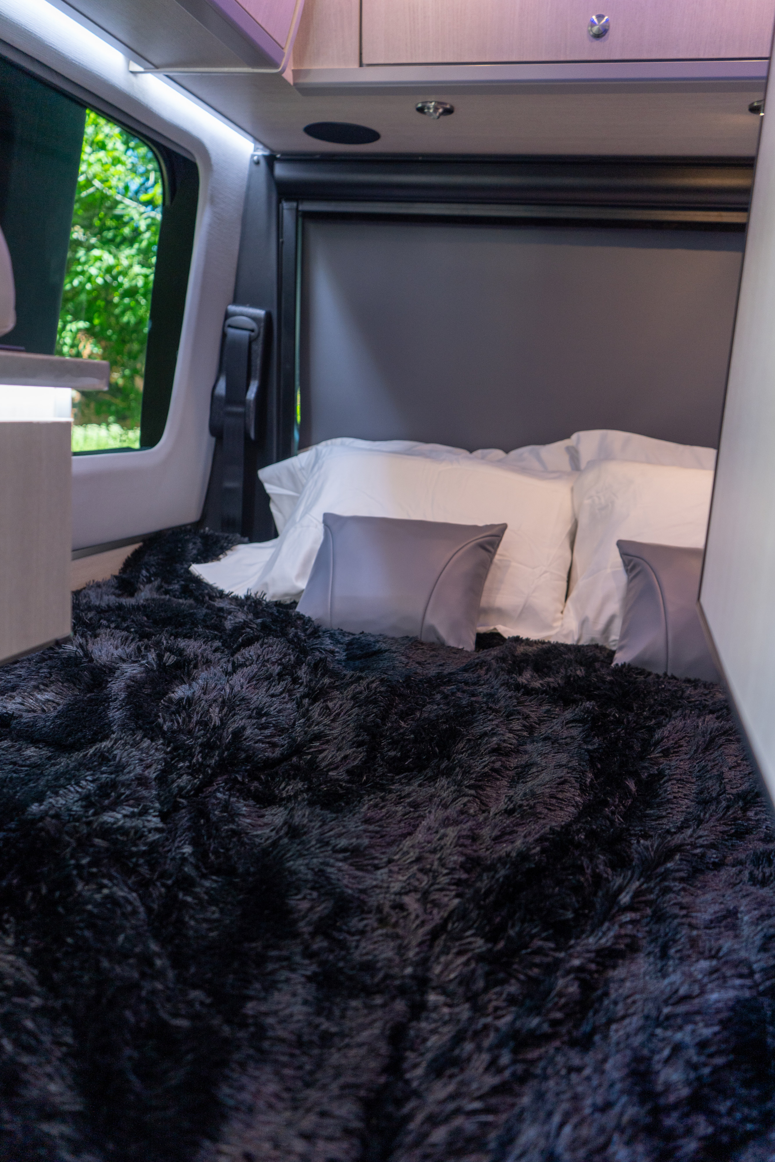 Cozy RV rental interior with a comfortable bed, featuring a plush black blanket, white pillows, and a window view of trees.. Grech RV Turismo Ion 2026