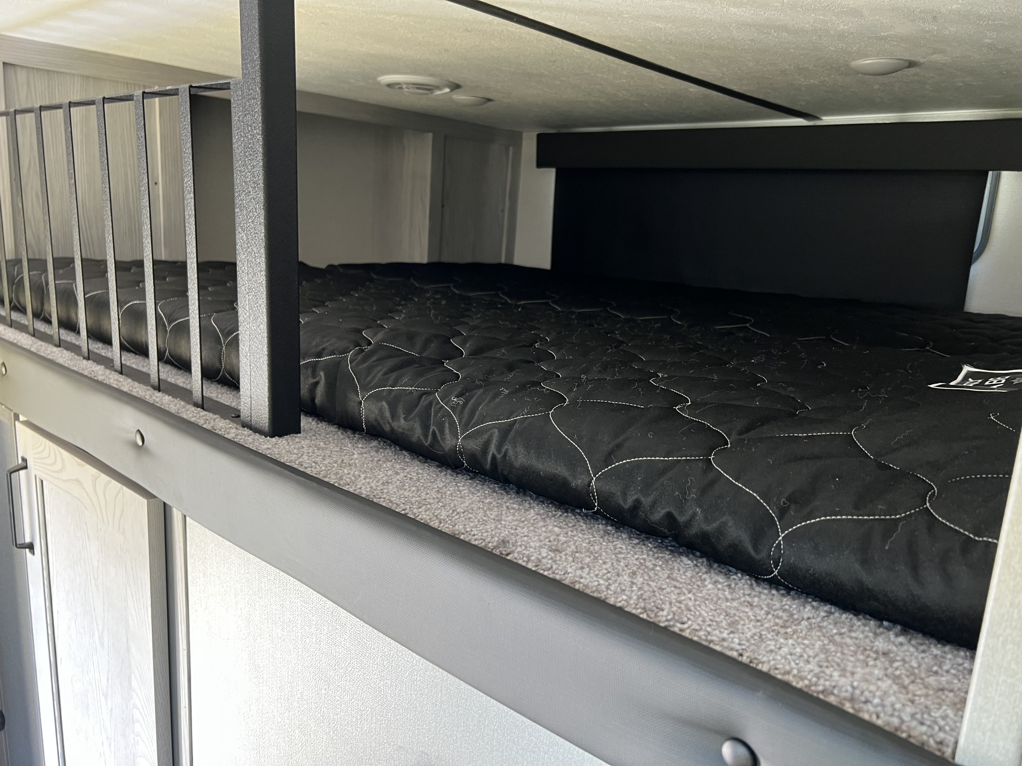 Close-up of a cozy RV rental bunk bed interior, featuring a black quilted mattress, metal safety rail, and carpeted sleeping area.. Keystone RV Montana 2022