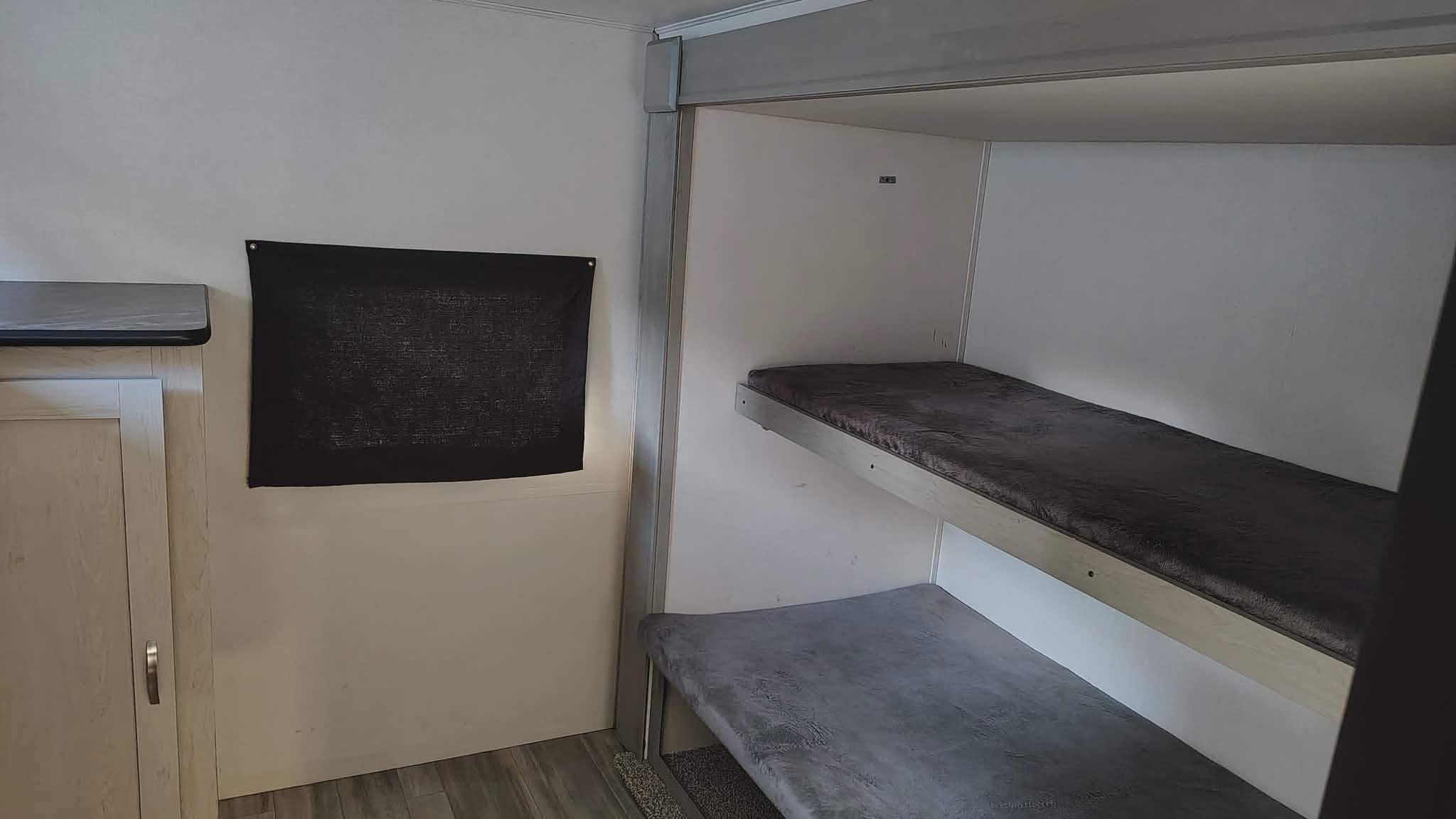 RV rental interior features bunk beds with mattresses, a small cabinet, and a dark fabric wall hanging for sleeping convenience.. Forest River Tracer 2022