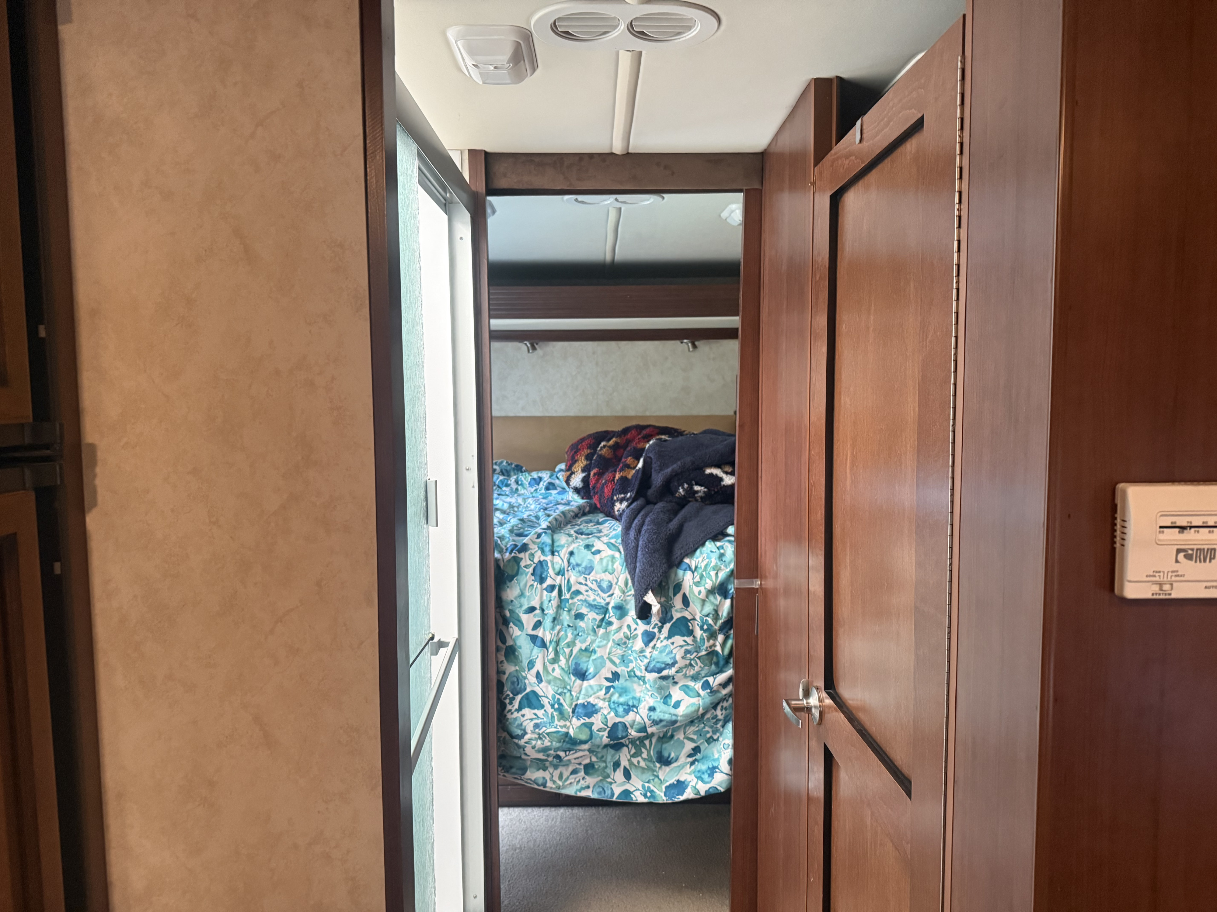 RV rental interior: A peek into a cozy bedroom with a floral comforter and blankets, seen through an open door. A thermostat is on the right wall.. Jamboree Jamboree Motorhome 2011