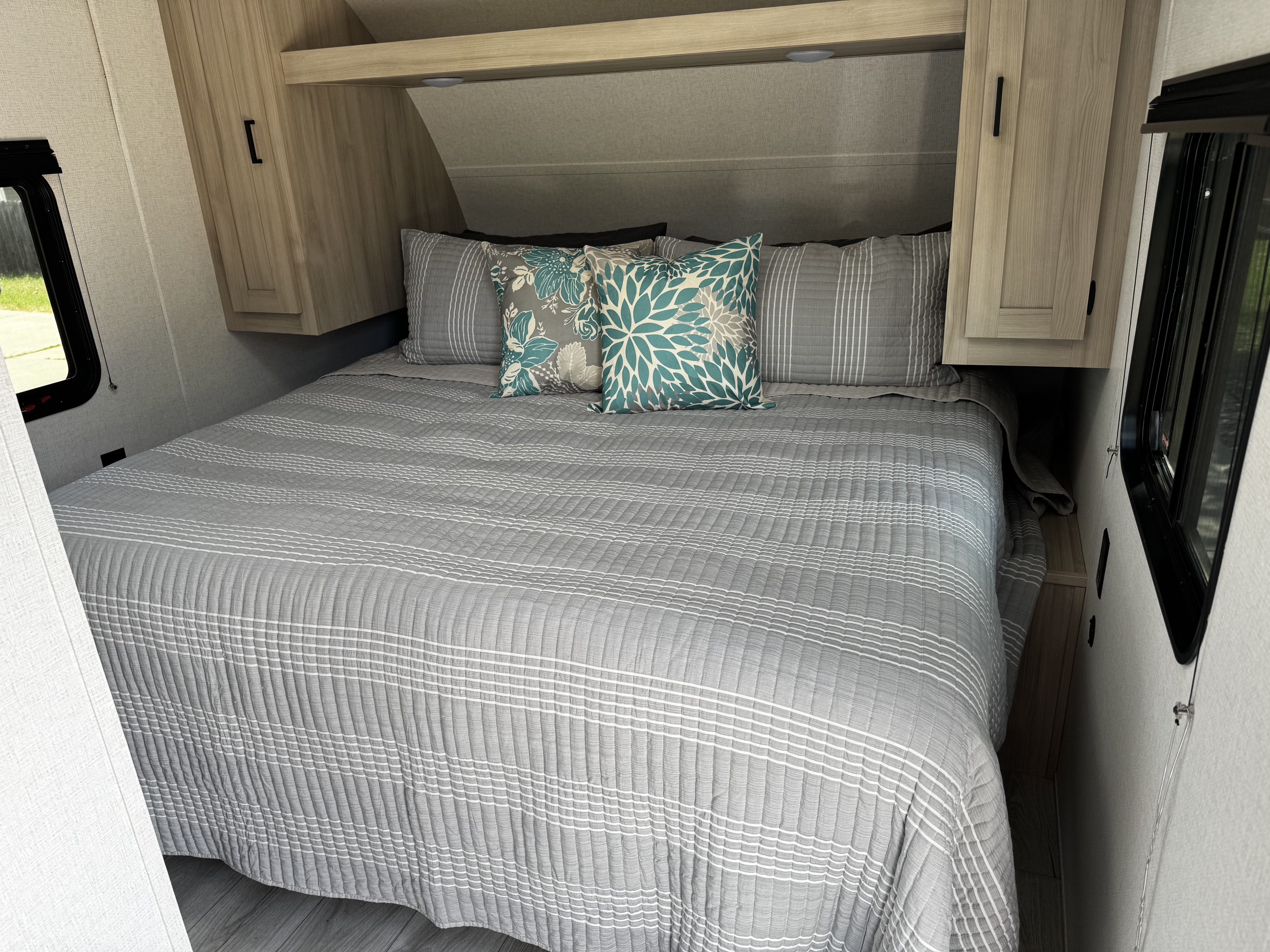 Cozy RV rental bedroom features a comfy grey bed with teal accents, overhead cabinets, and a side window, ideal for travel.. East to West Silver Lake 2024