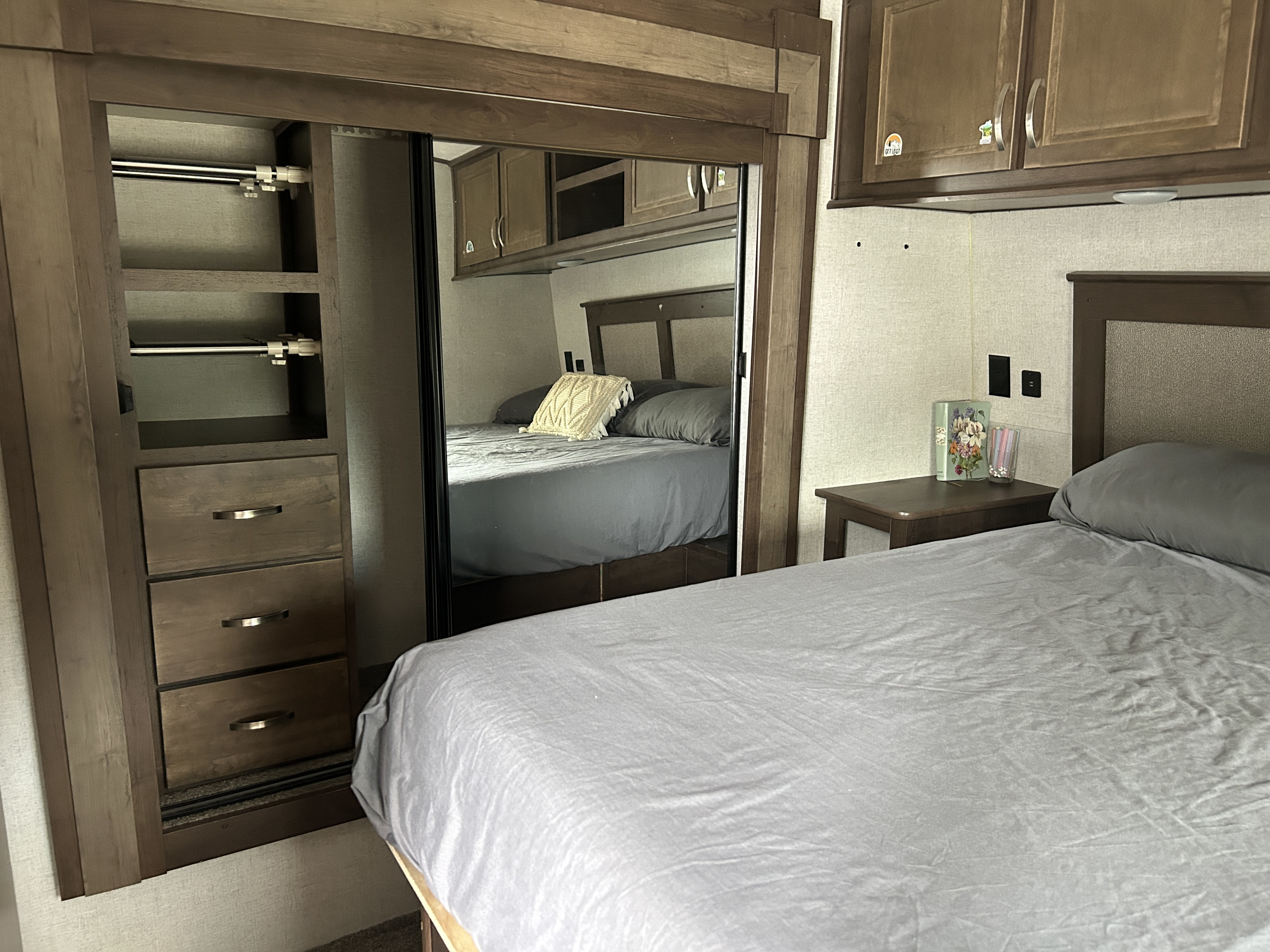 RV rental bedroom featuring a comfortable bed with grey linens, mirrored closet, built-in drawers, and overhead cabinets.. Highland Ridge RV Open Range 2020