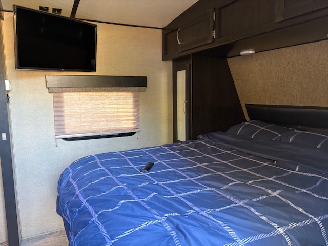 RV rental bedroom featuring a wall-mounted TV, large bed with blue plaid bedding, window with shade, and overhead cabinets.. Grand Design Imagine 2018
