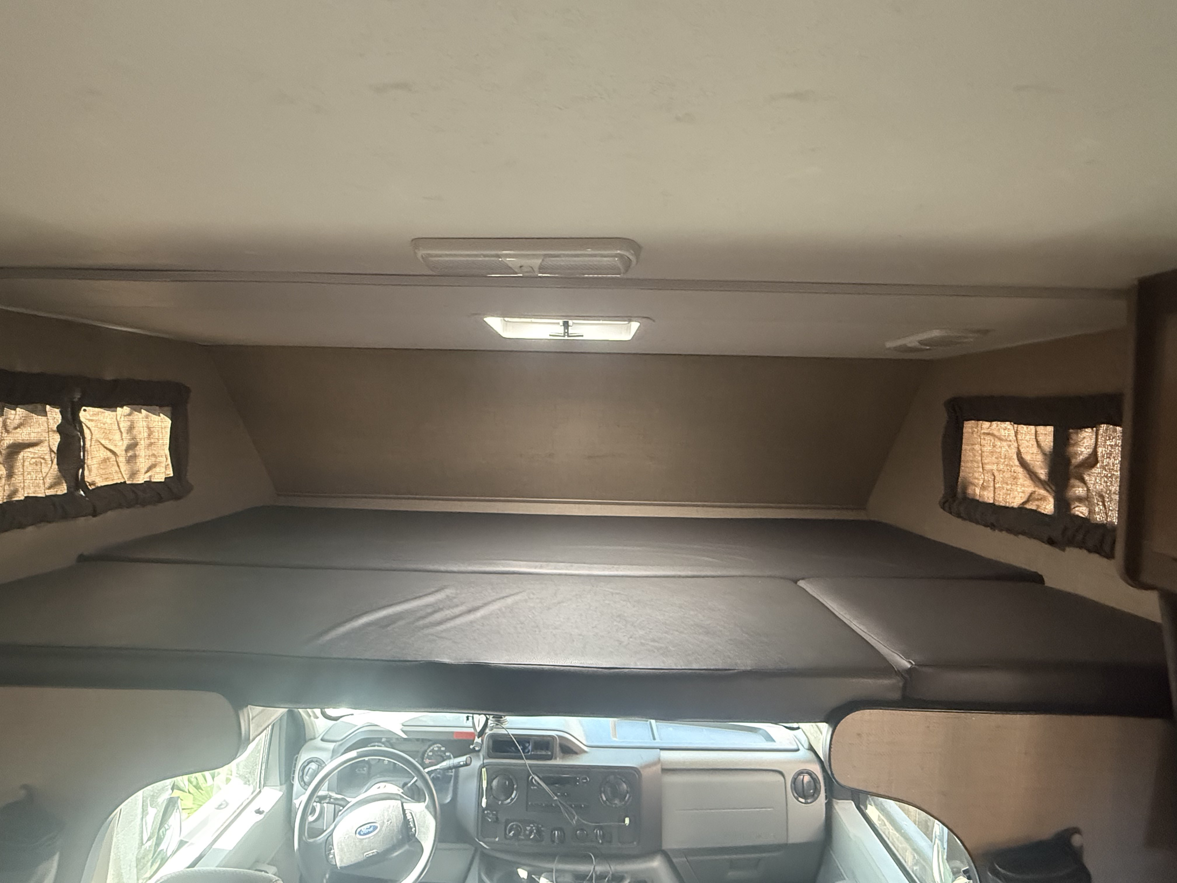 RV rental interior view: over-cab bunk bed, cockpit dashboard with steering wheel. Curtained windows on either side.. Thor Majestic 2019