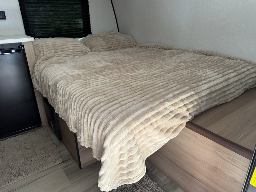RV rental bedroom featuring a soft, textured beige bed, adjacent mini-fridge, and window blinds.. Keystone RV Coleman Legacy 2026