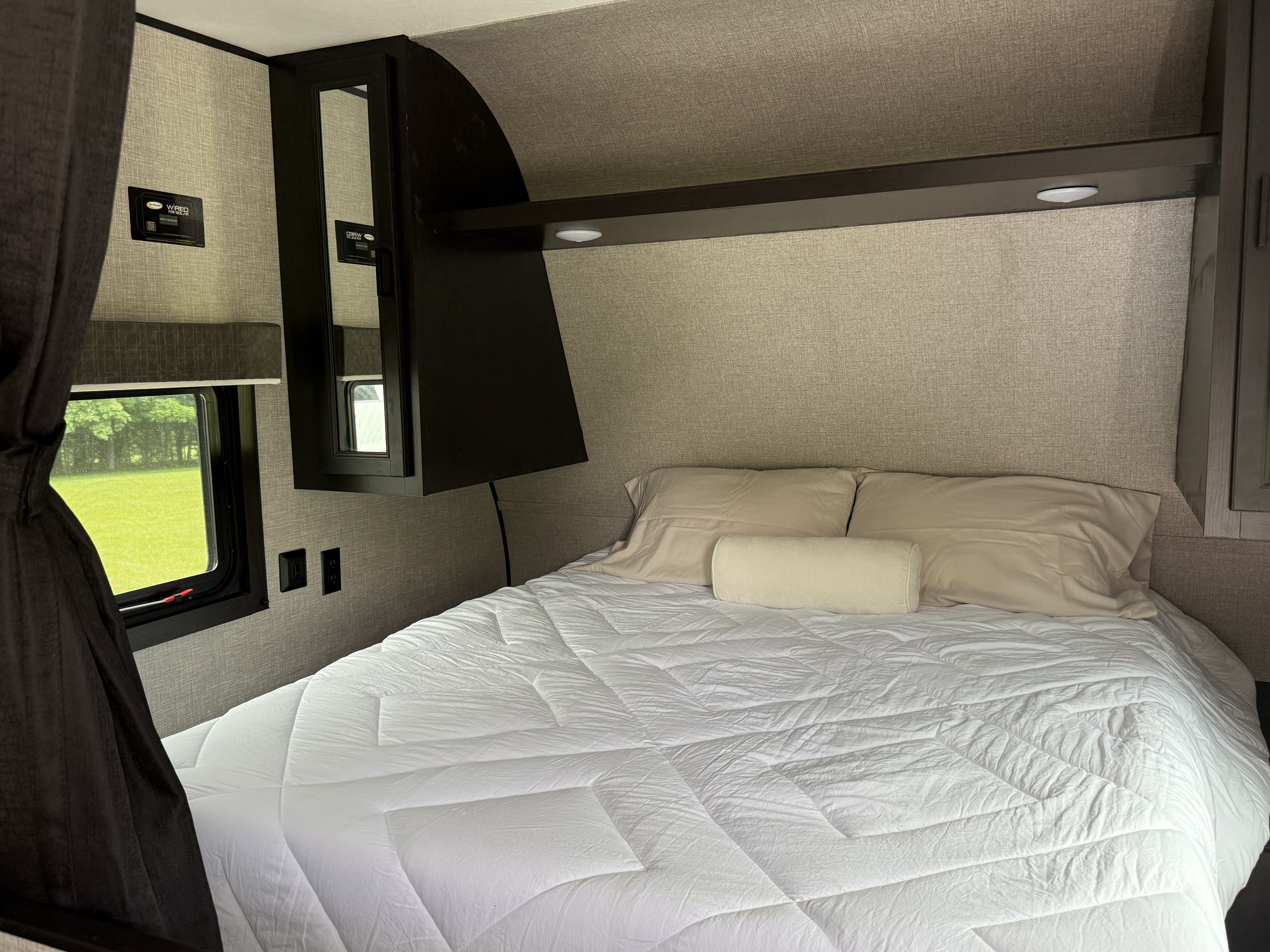 RV rental bedroom with a cozy bed, dark overhead storage, window to green scenery, and power outlets. Wired for solar.. Jayco Jay Flight SLX 2022