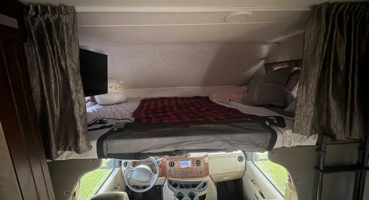 This RV rental interior features a cozy over-cab bed with a TV & safety net, overlooking the cockpit dashboard.. Forester 3051s 2018