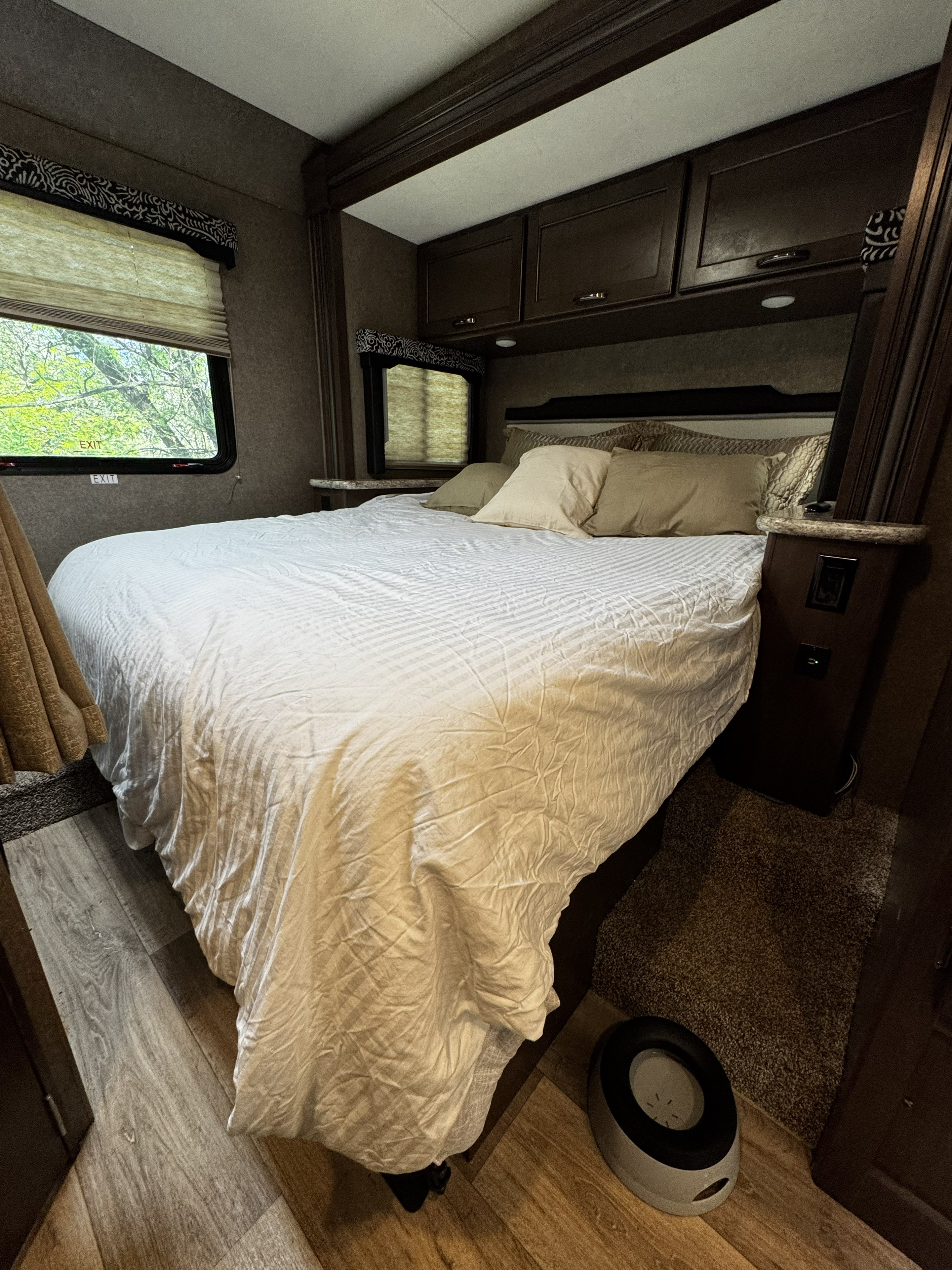 Cozy RV rental bedroom with a large bed, windows showing trees, overhead cabinets, and a pet bowl on the floor.. Thor Freedom Elite 2020
