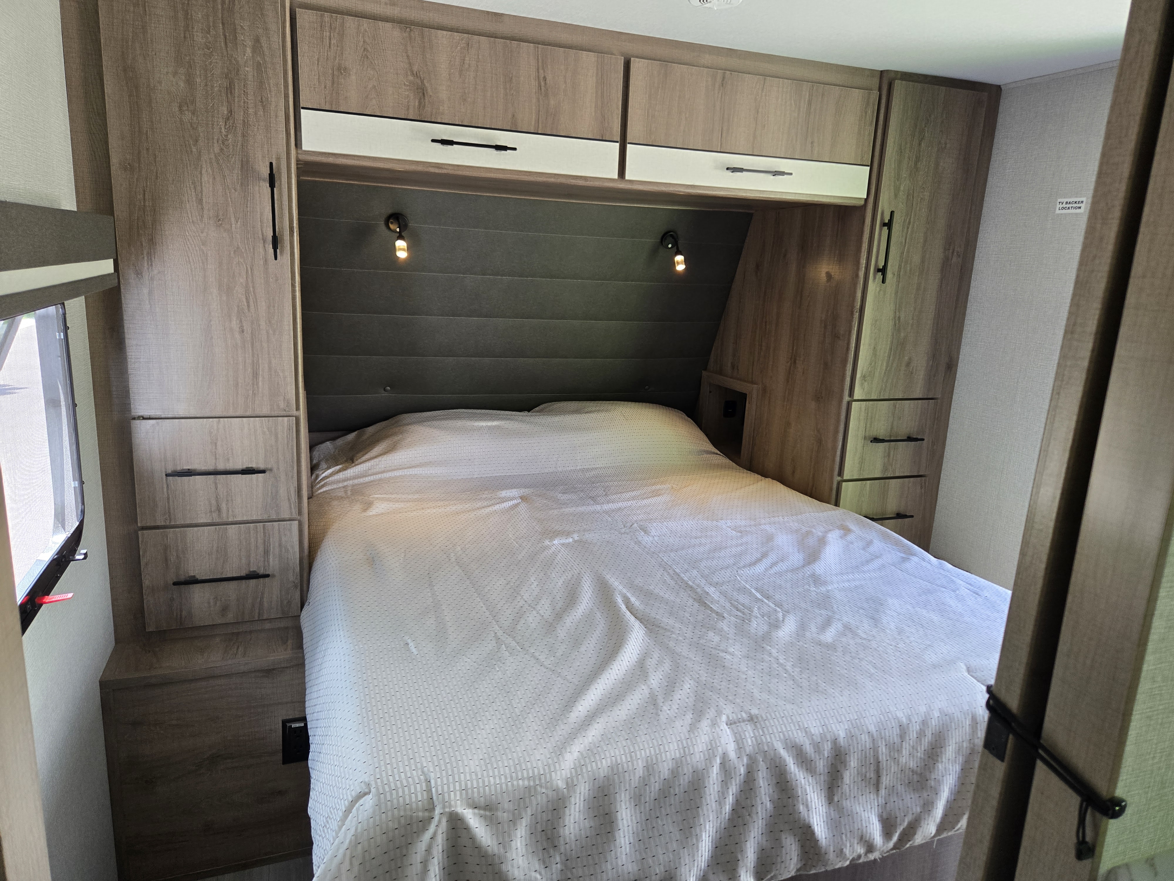 Inside a modern RV rental, a compact bedroom features a bed with overhead cabinets, bedside storage, and reading lights.. Grand Design Imagine XLS 2024
